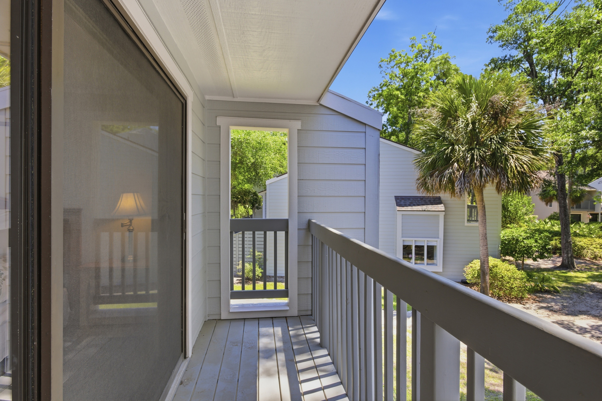 Striped shadows stretch across the cool grey floorboards of this private balcony. It is a quiet spot to breathe in the fresh air and gaze at the lush trees under a bright blue sky.