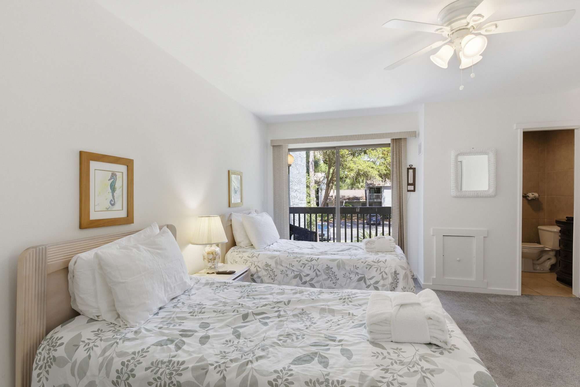 Floral linens and soft textures create a serene escape in this airy bedroom. Afternoon rays stream through glass doors, highlighting the clean, simple finishes and a private balcony view of the shaded greenery outside.
