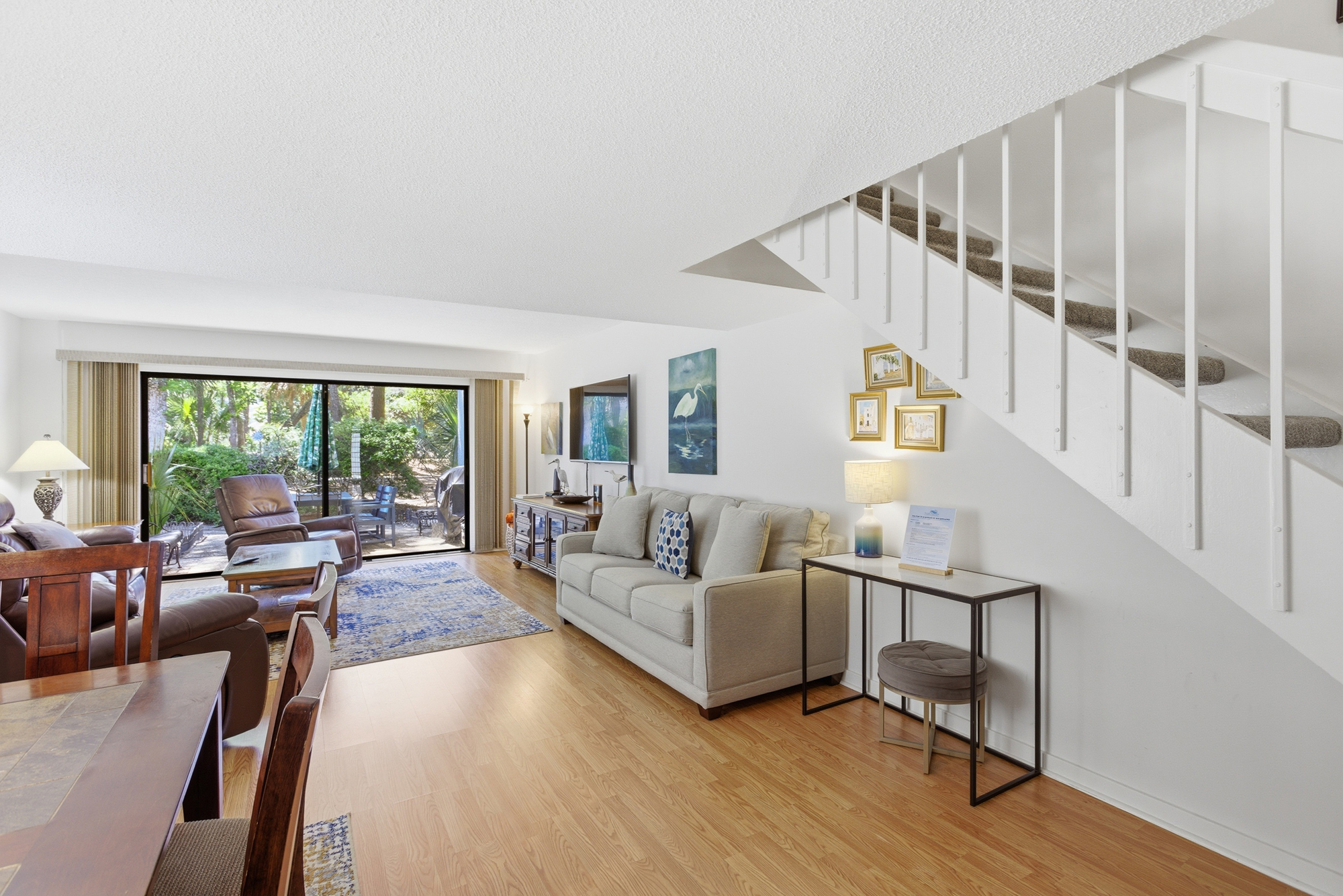 Honey-toned floors glow beneath a soaring white staircase, leading your eye toward a cozy seating area and lush garden views. Neutral fabrics and rich wood accents create a peaceful atmosphere for quiet afternoons and coastal dreaming.