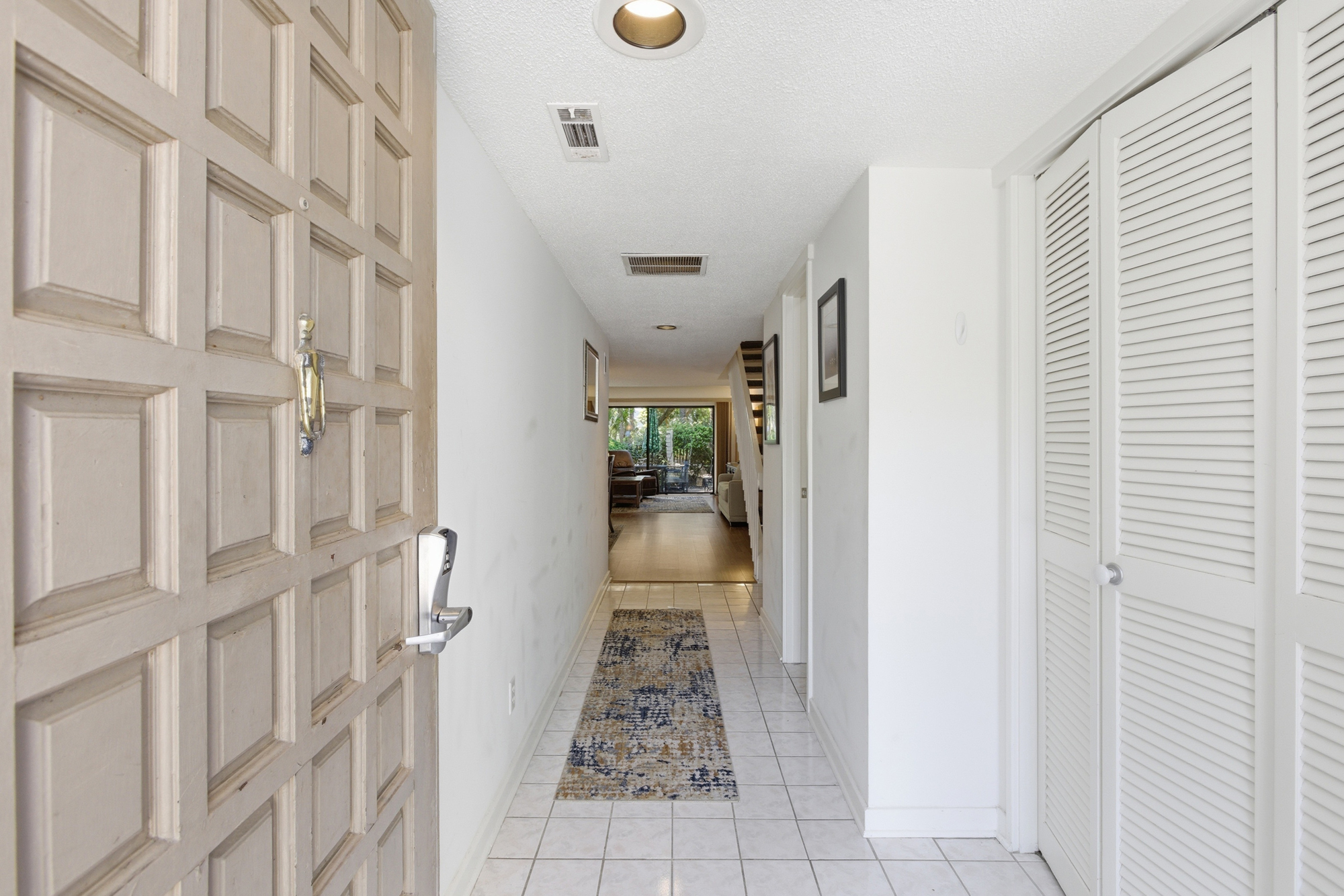 Carved wood paneling on the front door opens into a bright, airy hallway where cool tiles and a patterned runner lead your eye toward the lush greenery of the patio. Soft overhead beams illuminate the path into this crisp, serene living space.