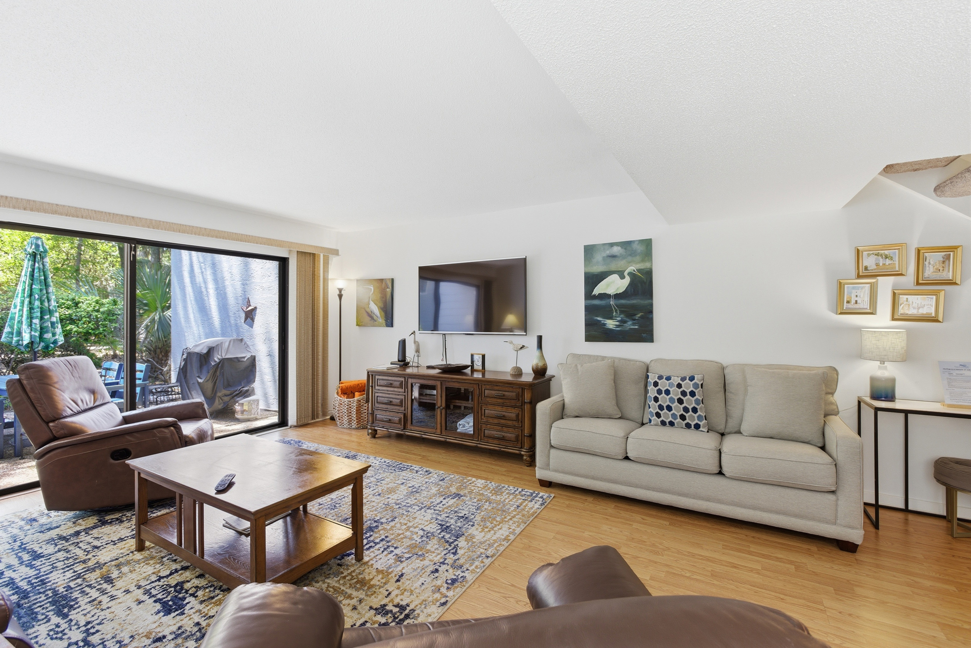 Natural illumination pours through sliding glass doors, illuminating the oak floors and plush seating of this airy lounge. Earthy wood tones and coastal art create a grounded, serene atmosphere perfect for quiet afternoons by the patio.