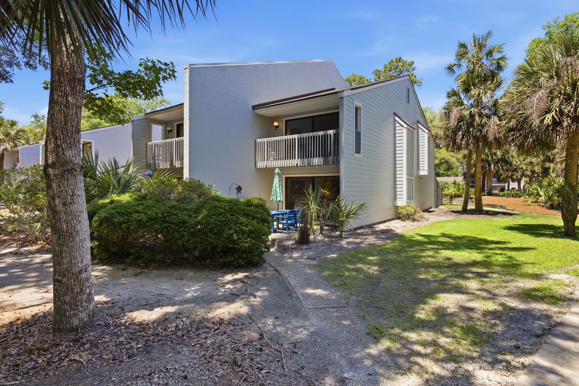Sandy paths and lush greenery lead to your quiet balcony, where soft shadows dance across the modern facade. This peaceful corner offers a bright, airy space to sit outside and breathe in the salty air under a clear blue sky.
