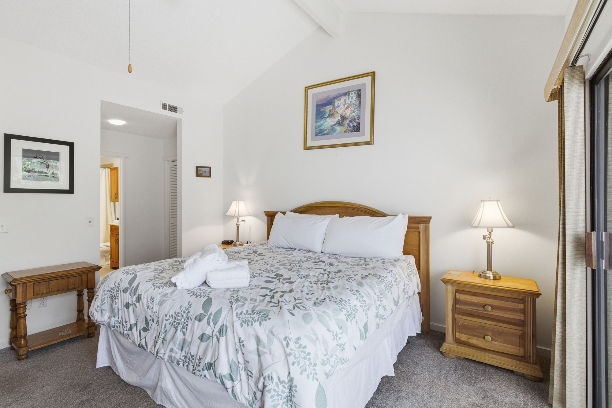 Golden wood grains and botanical prints bring a quiet, organic charm to this airy bedroom. Soft lamps cast a warm glow across the vaulted ceilings and fresh linens, while sliding doors suggest a seamless connection to the outdoors.