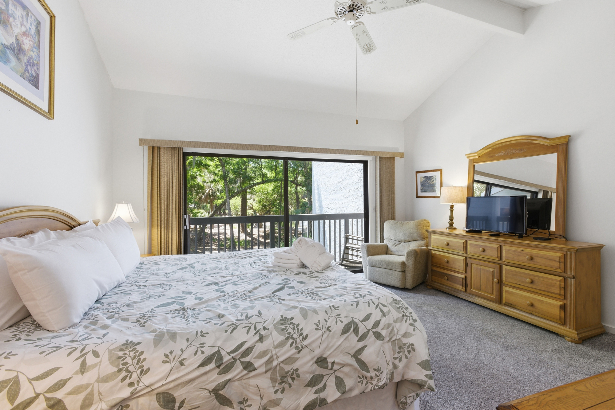 Leafy views frame this airy primary suite, where vaulted ceilings and warm oak furniture create a calm atmosphere. Crisp linens and a plush armchair invite quiet afternoons spent watching the trees sway through the sliding glass doors.