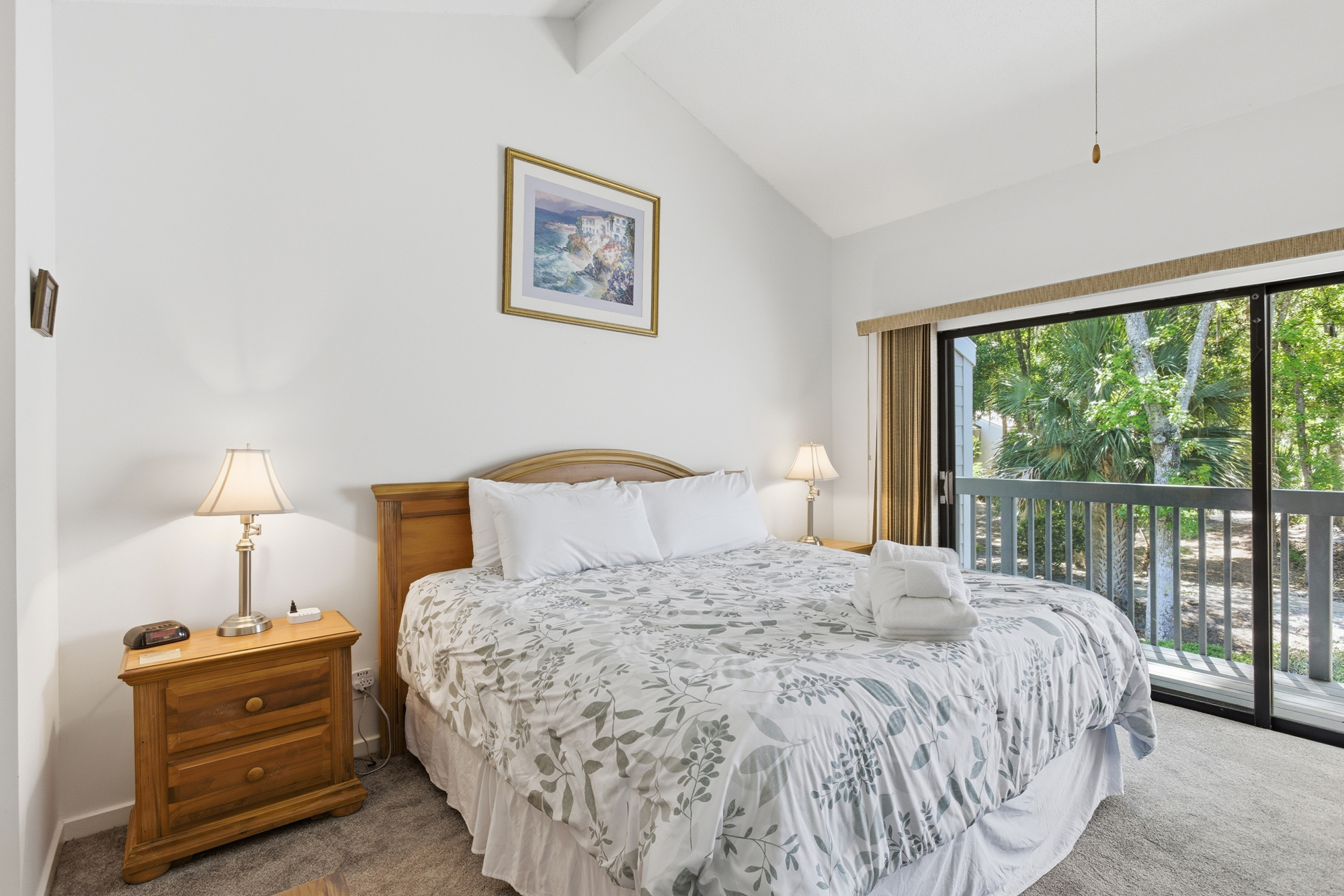 Tall vaulted ceilings and sliding glass doors create an airy, open sanctuary where leafy views bring the outdoors inside. Soft bedside lamps cast a golden glow over the warm wood grains and botanical prints for a peaceful night of rest.
