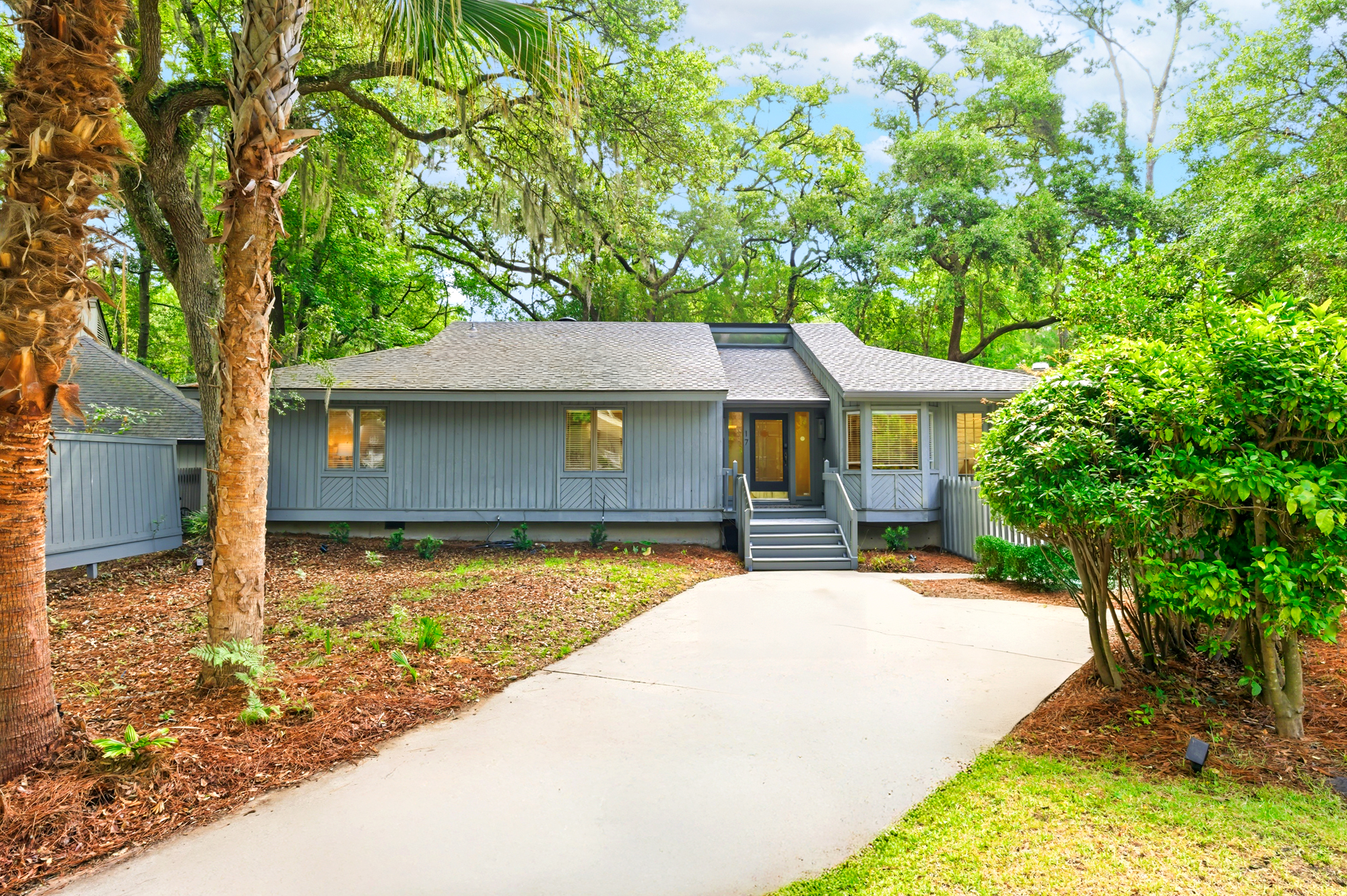 Ancient oaks and swaying palms frame this modern cottage, where soft grey siding meets the lush green of a coastal forest. Golden light glows from within, promising a quiet and calm stay under the sprawling southern canopy.