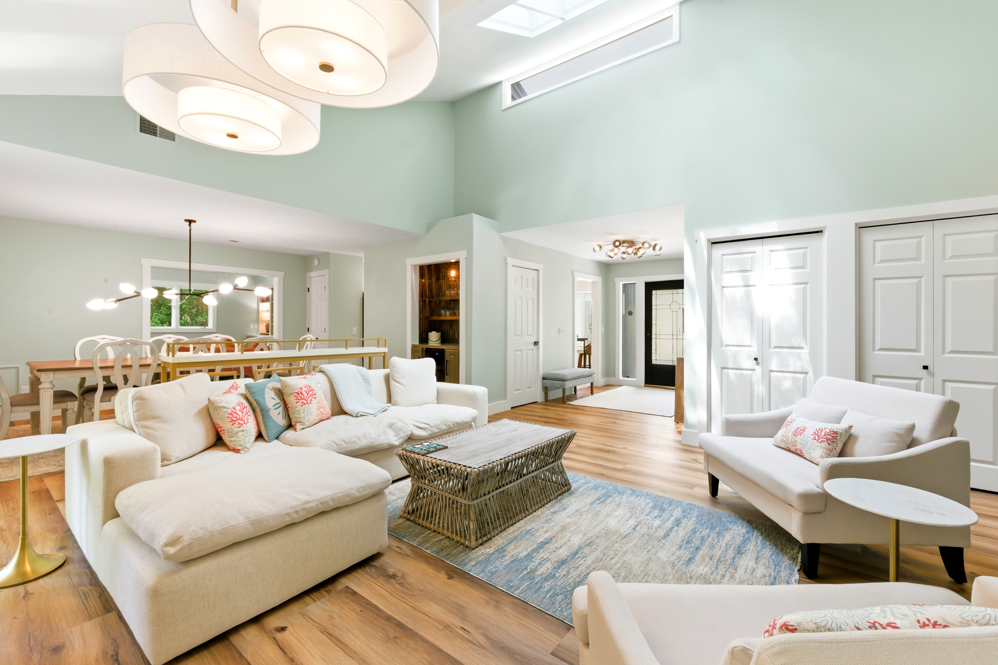 Lofty seafoam walls and vaulted ceilings create an airy backdrop for this open living area. Plush ivory seating and a textured wicker table rest upon wide oak planks, illuminated by a soft daytime glow from the clerestory windows above.
