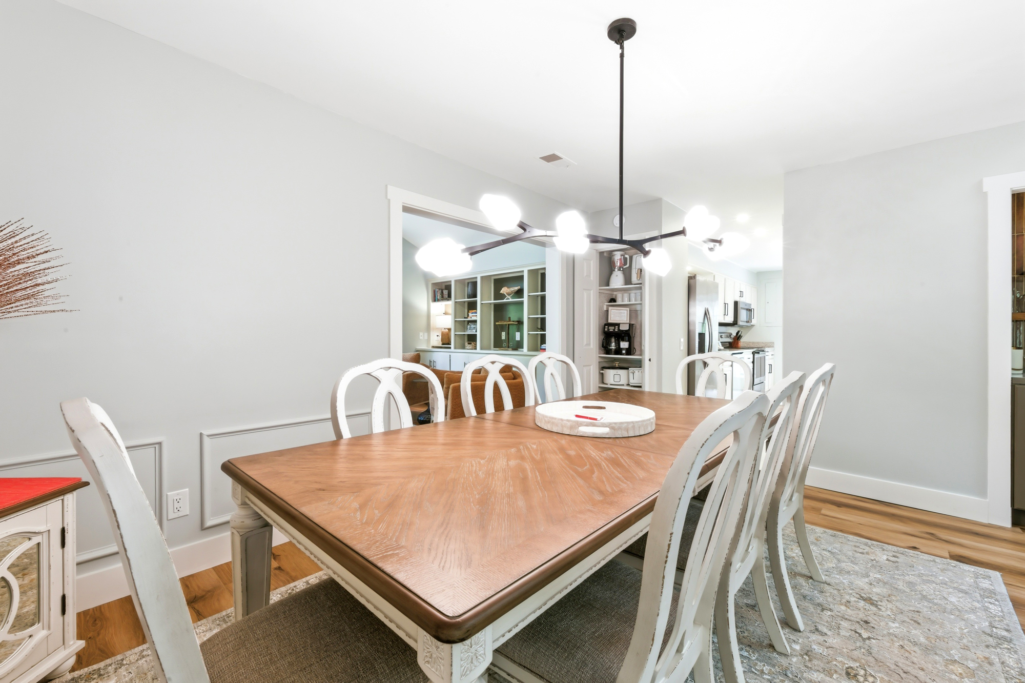 Warm wood grains and elegant white seating anchor this airy dining space, illuminated by a sculptural modern chandelier. Soft gray walls and open sightlines create a calm, social atmosphere perfect for long morning coffees and shared meals.