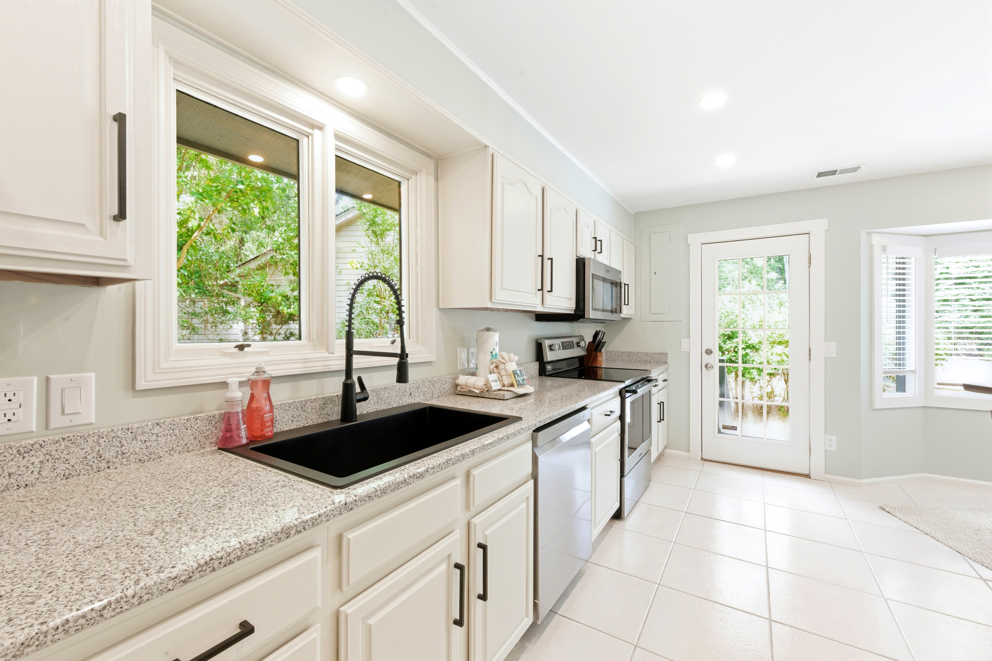 Granite countertops and crisp white cabinetry create a clean, airy kitchen workspace. Afternoon glow pours through the broad window, framing a view of lush garden greenery while you prepare your favorite morning coffee.