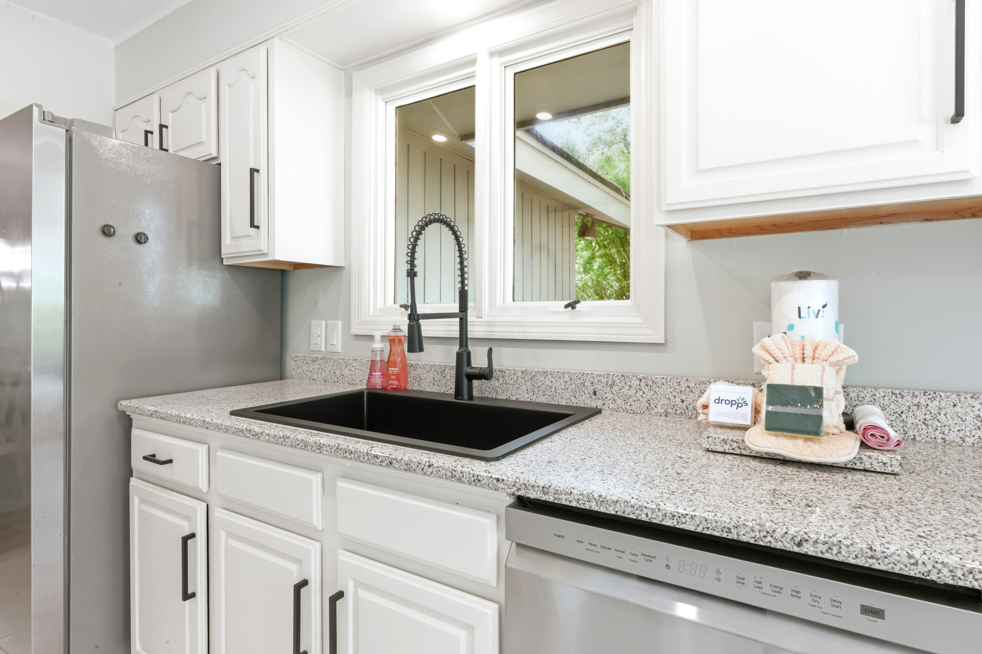 Crisp white cabinetry and speckled stone counters frame this modern kitchen corner, where a sleek black faucet stands ready for your morning coffee prep. Natural light pours through the window, highlighting the clean, thoughtful details.