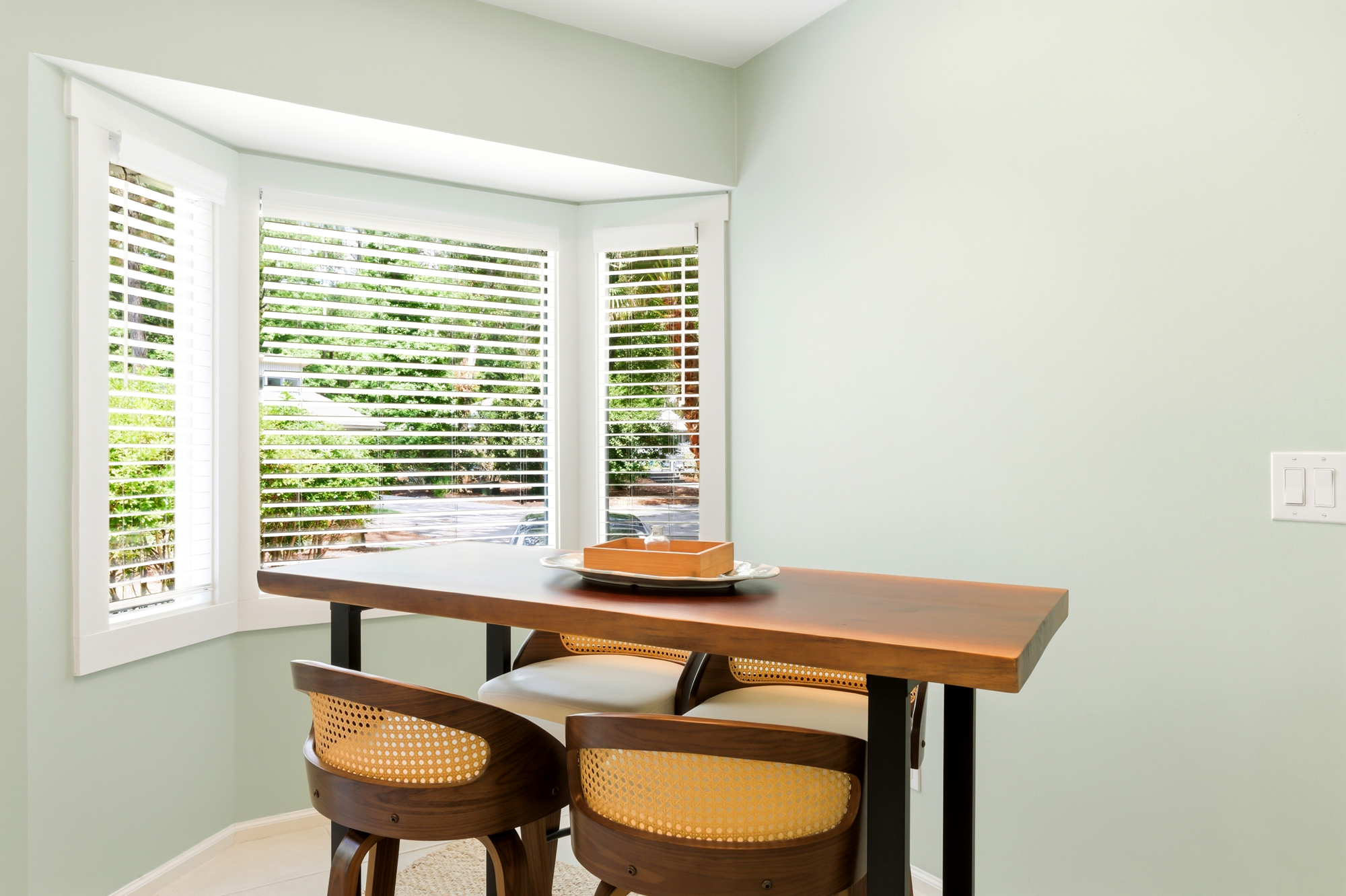 Rich timber grains and cane textures anchor this breakfast nook, where a bay window filters a soft afternoon glow over the sage walls. It is a quiet corner for morning coffee or intimate conversation.