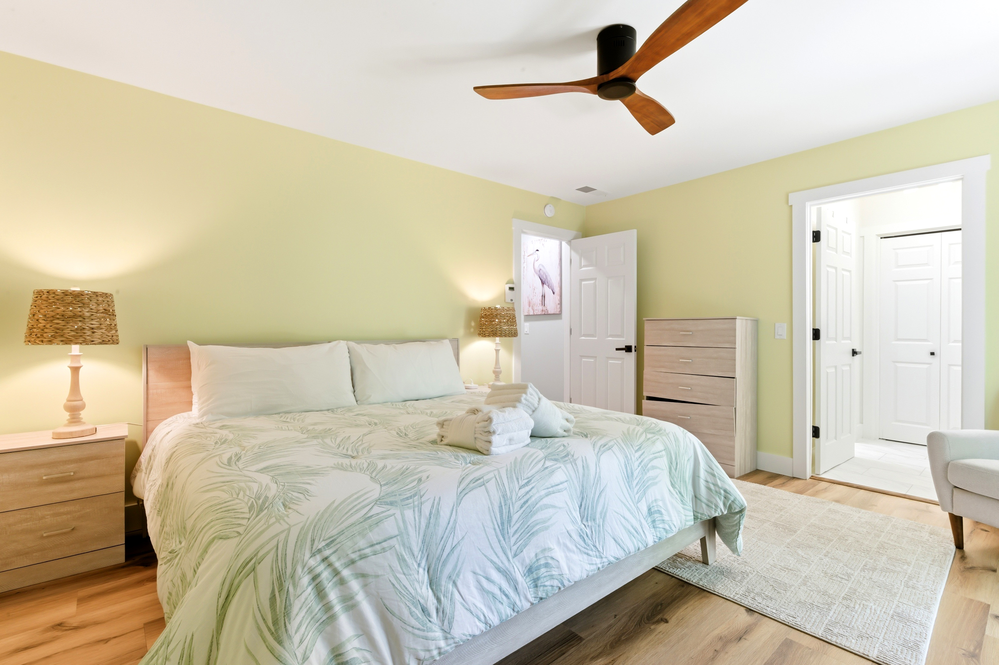 Palm-leaf patterns and honey-toned wood floors create a serene, airy atmosphere in this quiet bedroom. Natural textures from the rattan lamps cast a gentle glow over the plush linens, perfect for a peaceful night of rest.