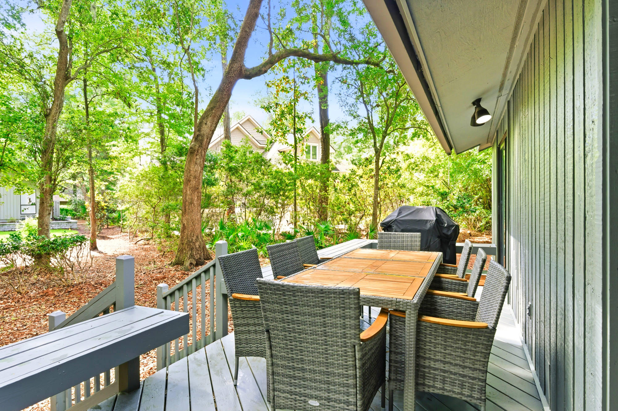 Woven chairs and a warm wood table anchor this breezy deck, creating a perfect spot for alfresco meals. Tall trees filter the afternoon glow, surrounding the space with lush greenery and a peaceful, airy atmosphere.