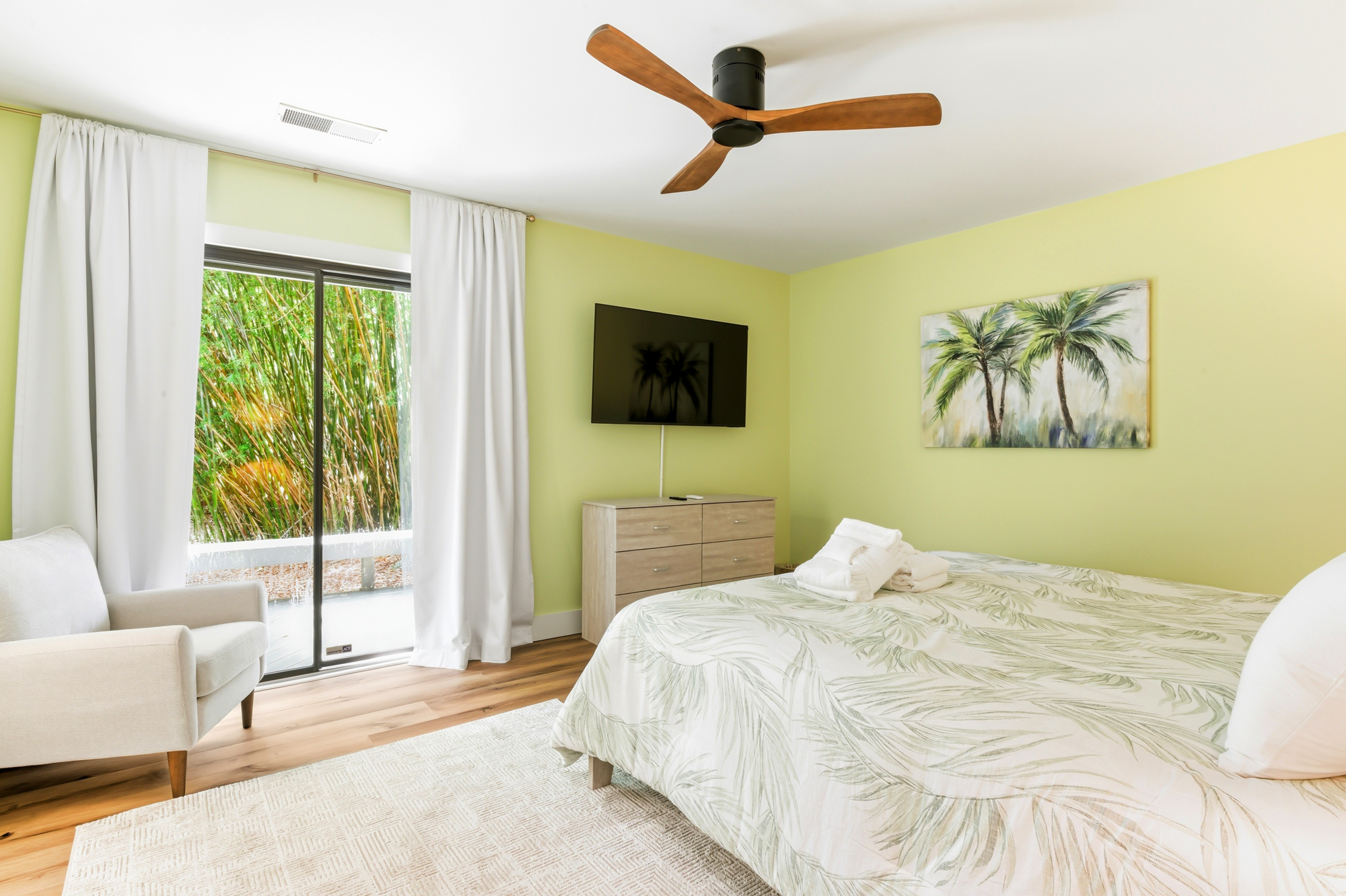 Pale lime walls and botanical bedding create a serene indoor garden feel, framed by a view of lush greenery through sliding glass doors. Natural wood tones and a gentle breeze from the sleek ceiling fan complete this airy, sun-drenched sanctuary.
