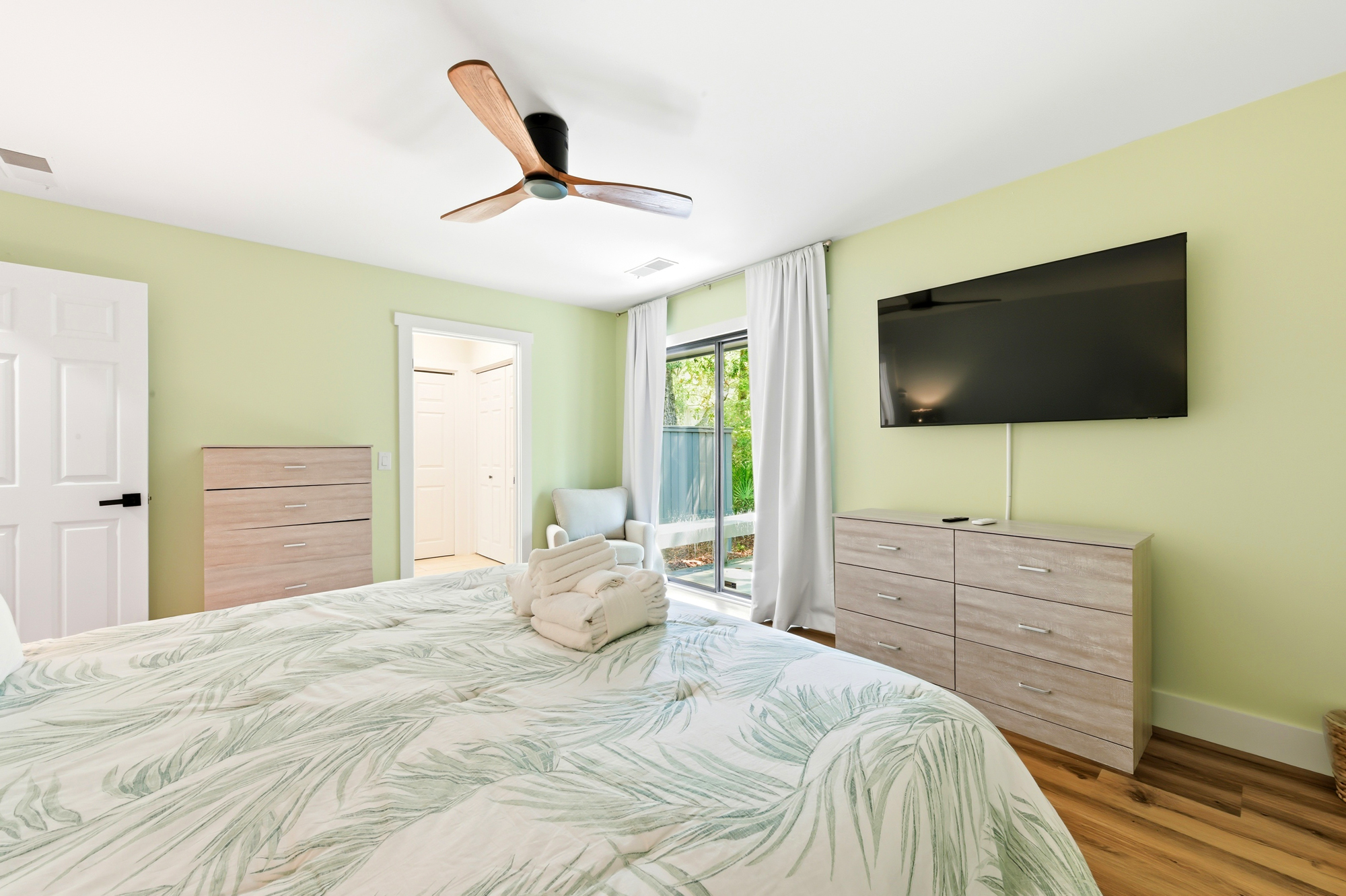 Soft green walls and botanical linens create a tranquil backdrop for slow mornings. Natural wood accents and a sleek ceiling fan bring a sense of organic calm to this airy bedroom, which opens directly via glass doors to the lush greenery outside.