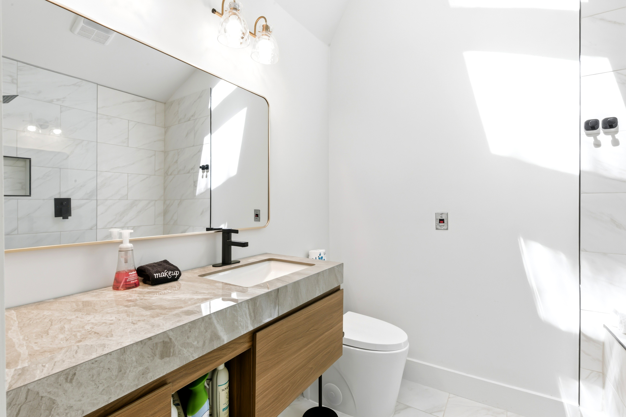 Marble textures and sleek wood cabinetry create a serene atmosphere in this airy bathroom. Natural afternoon glow reflects off the polished surfaces, highlighting the modern fixtures and spacious mirror for a refreshing start to your day.