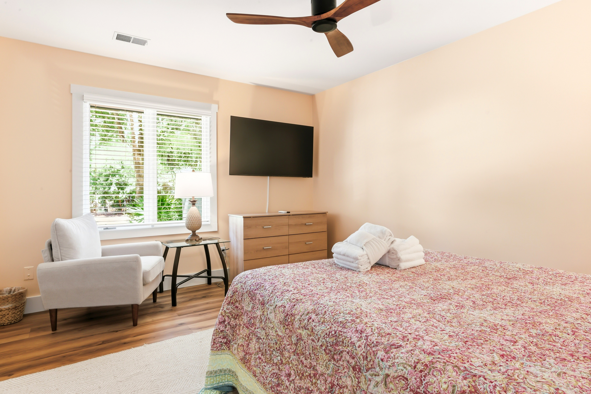 Peach-hued walls create a calming glow in this quiet bedroom, where a plush armchair waits by the window. Wood grains and botanical patterns ground the space, offering a serene corner for slow mornings and restful nights.