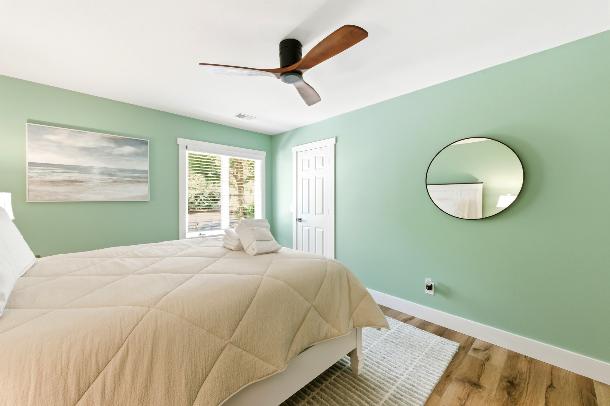 Mint walls and a sculpted wood fan create a serene, airy atmosphere in this quiet bedroom. Natural light spills across the soft duvet and honey-toned floors, offering a peaceful spot to rest after a day of travel.