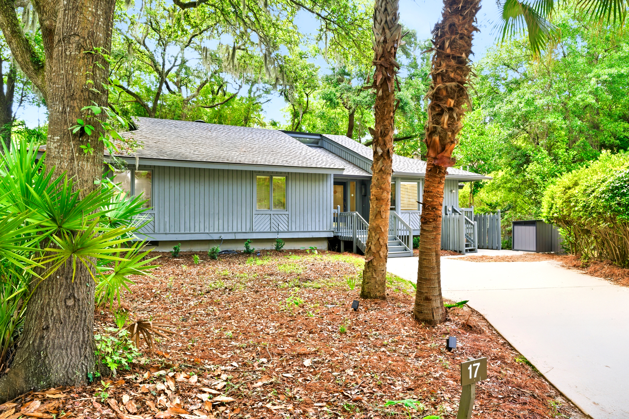 Spanish moss drapes from towering oaks to frame this coastal cottage, where soft gray siding harmonizes with the surrounding forest. Tall palms stand guard over the winding path, leading you into a sun-drenched sanctuary of peace and privacy.