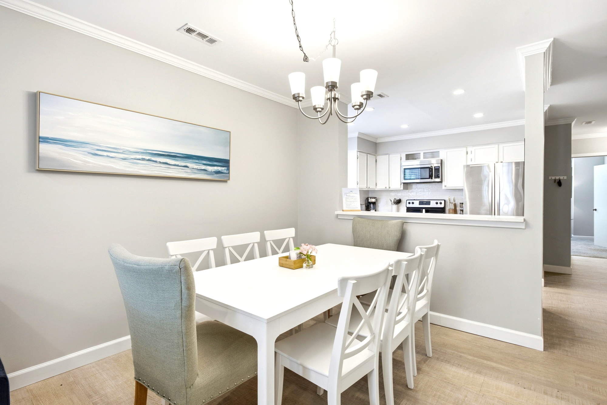Crisp white furniture and coastal art create a serene setting for morning coffee or shared meals. This airy dining space flows seamlessly into a modern kitchen, glowing under the gentle radiance of a classic chandelier.