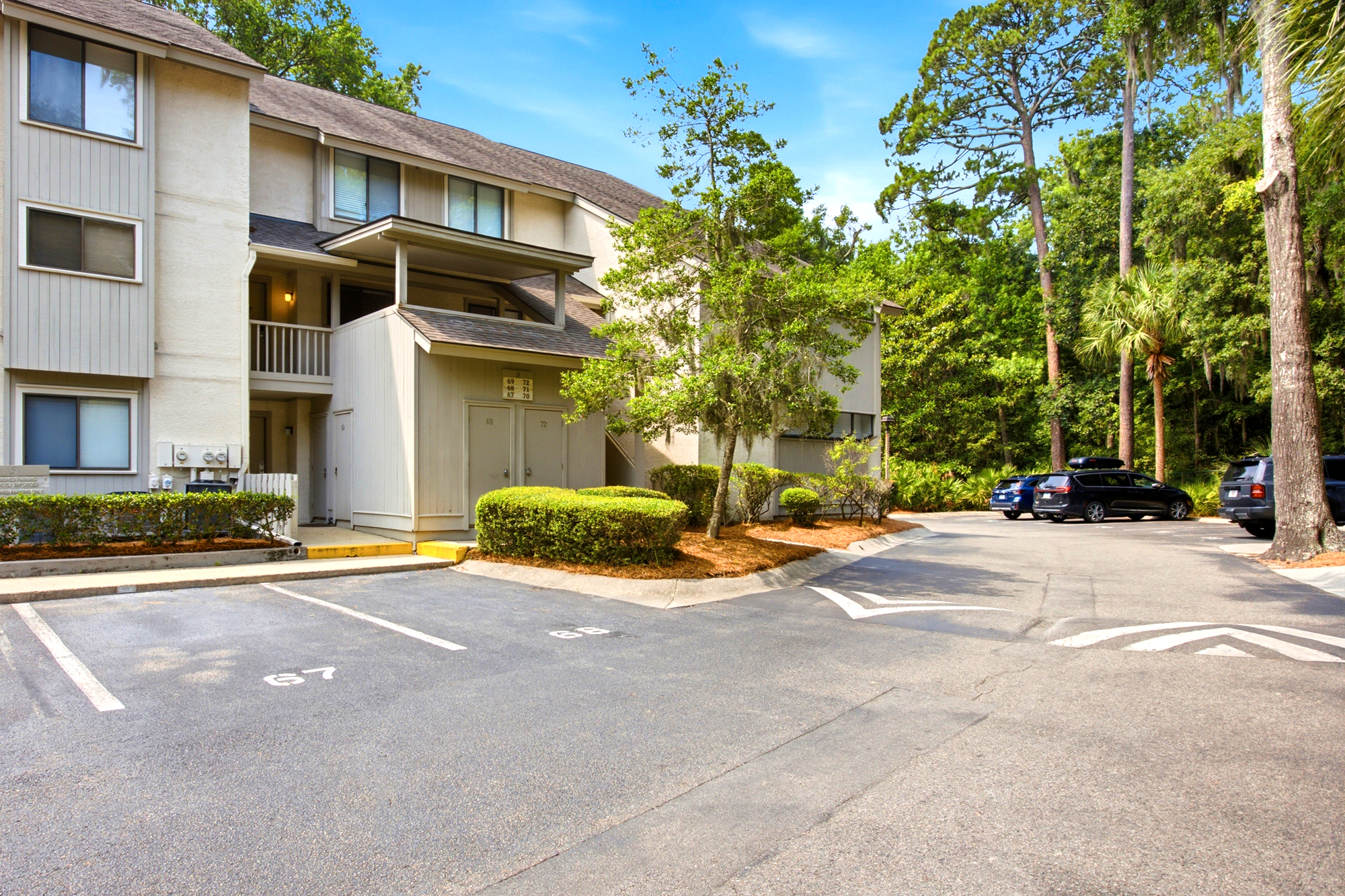 Tall pines and manicured hedges frame this quiet coastal residence, where soft afternoon glow hits the neutral siding. Ample parking and a private balcony offer a seamless transition between the airy interior and the lush, shaded surroundings.