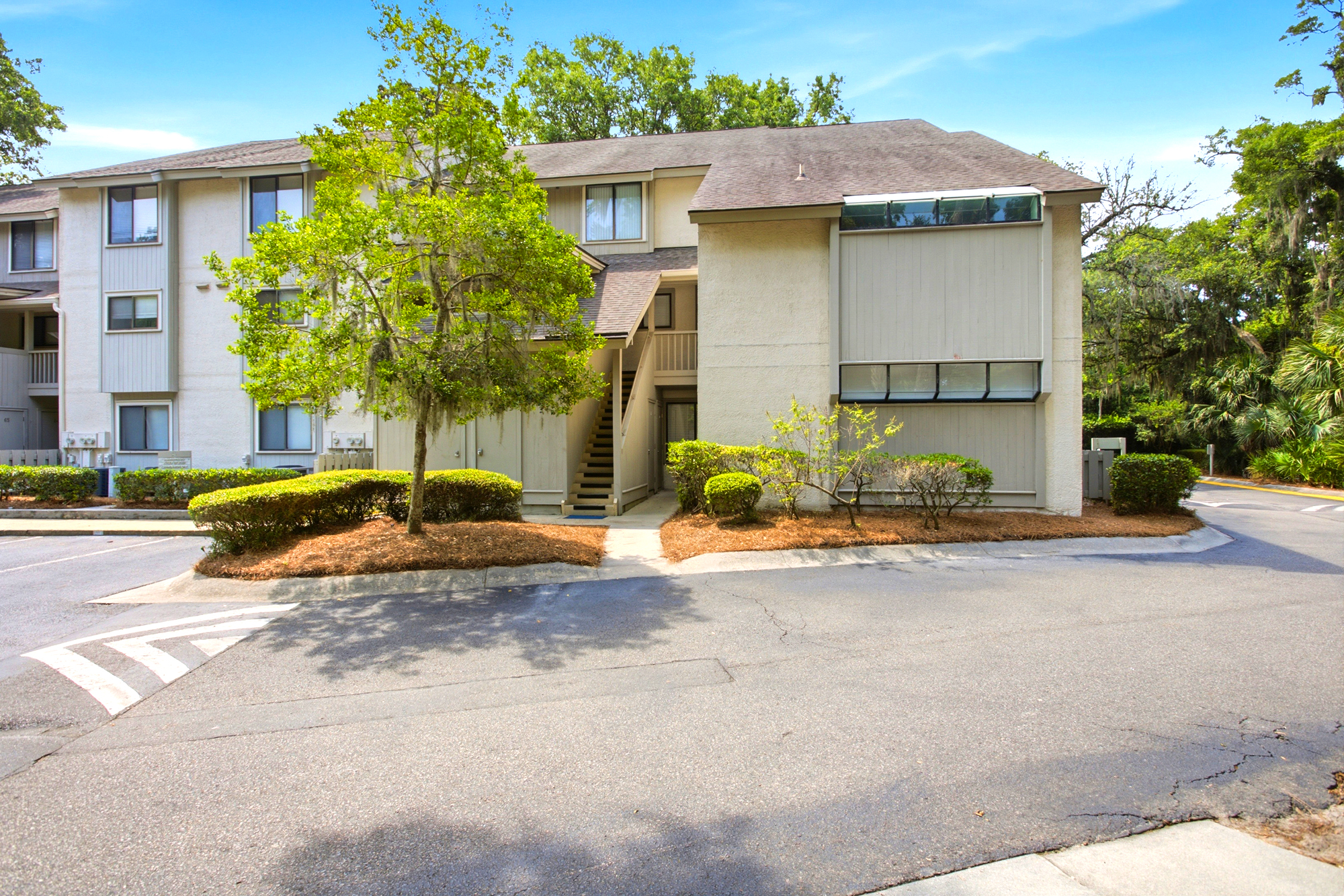 Leafy oak trees and manicured hedges frame this quiet cedar-shingled exterior, offering a peaceful first impression. Bright blue skies and soft afternoon shadows highlight the clean architectural lines of your coastal home.