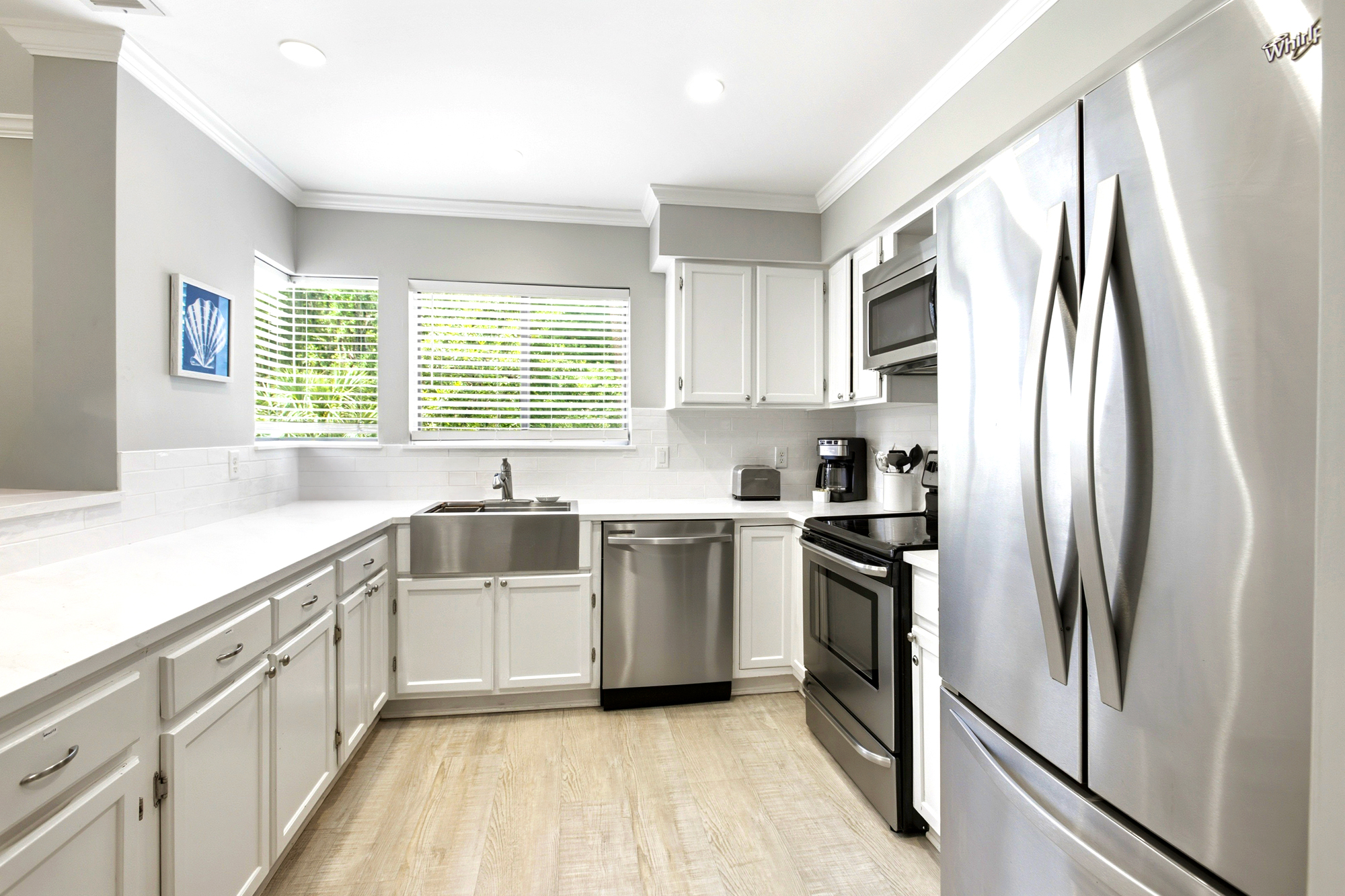 Polished stainless steel and crisp white cabinetry create a luminous, modern space for morning coffee. Natural light filters through the blinds, highlighting the sleek apron sink and airy layout designed for effortless coastal cooking.