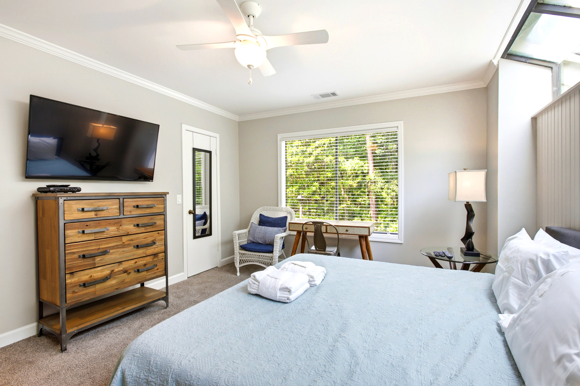 Natural wood textures and coastal blue linens create a serene atmosphere in this airy bedroom. An afternoon glow filters through the blinds, highlighting the cozy wicker armchair and functional workspace perfect for quiet reflection.