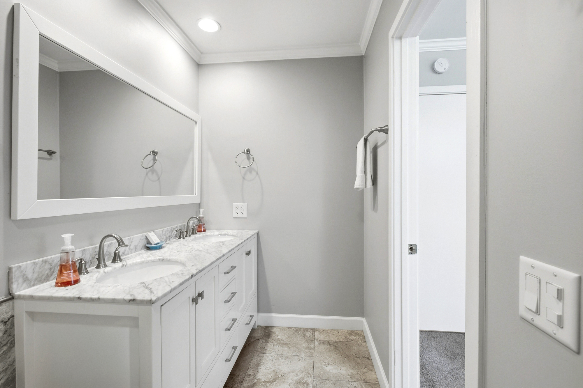 Marble surfaces and cool gray tones create a serene atmosphere in this airy bathroom. A double vanity with sleek fixtures offers ample space for morning routines under the bright, even glow of the recessed lighting.
