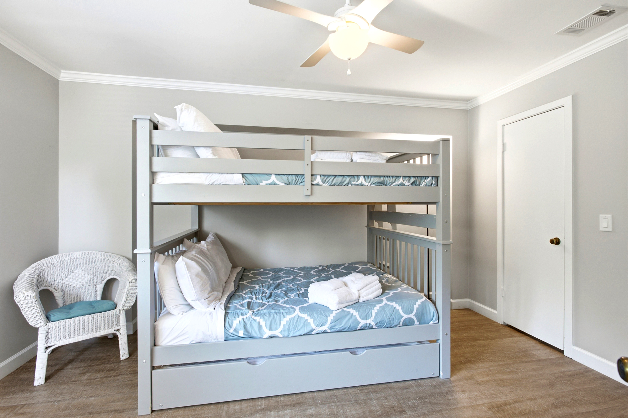 Slate gray bunk beds paired with patterned azure linens create a cheerful, bright nook for guests. A white wicker chair and honey-toned floors add a cozy, coastal touch to this clean and airy bedroom.