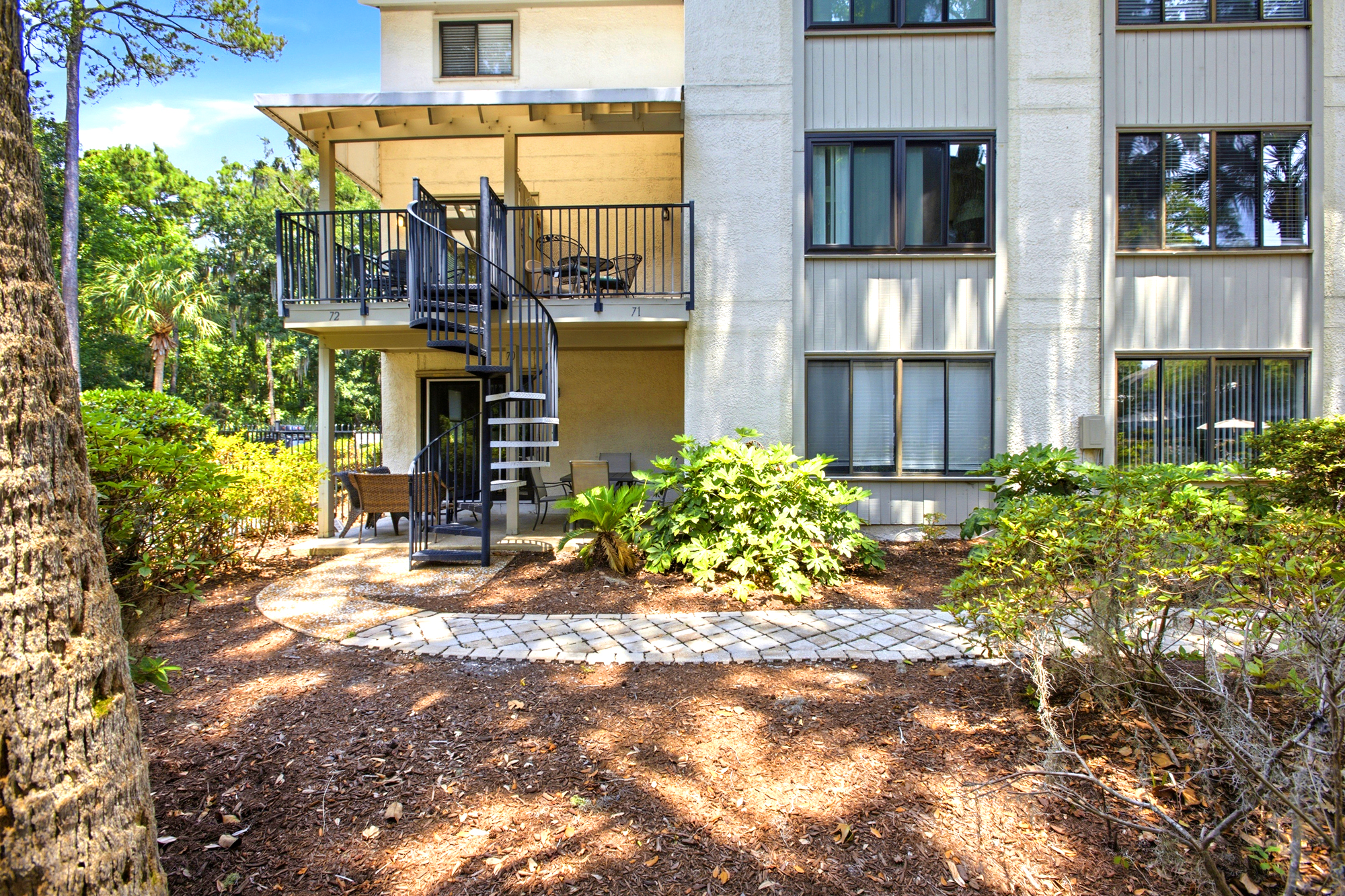 Ironwork spirals upward to a private balcony, connecting the lush garden path to your elevated outdoor sanctuary. Golden rays filter through the trees, highlighting the earthy textures and quiet charm of this secluded coastal escape.