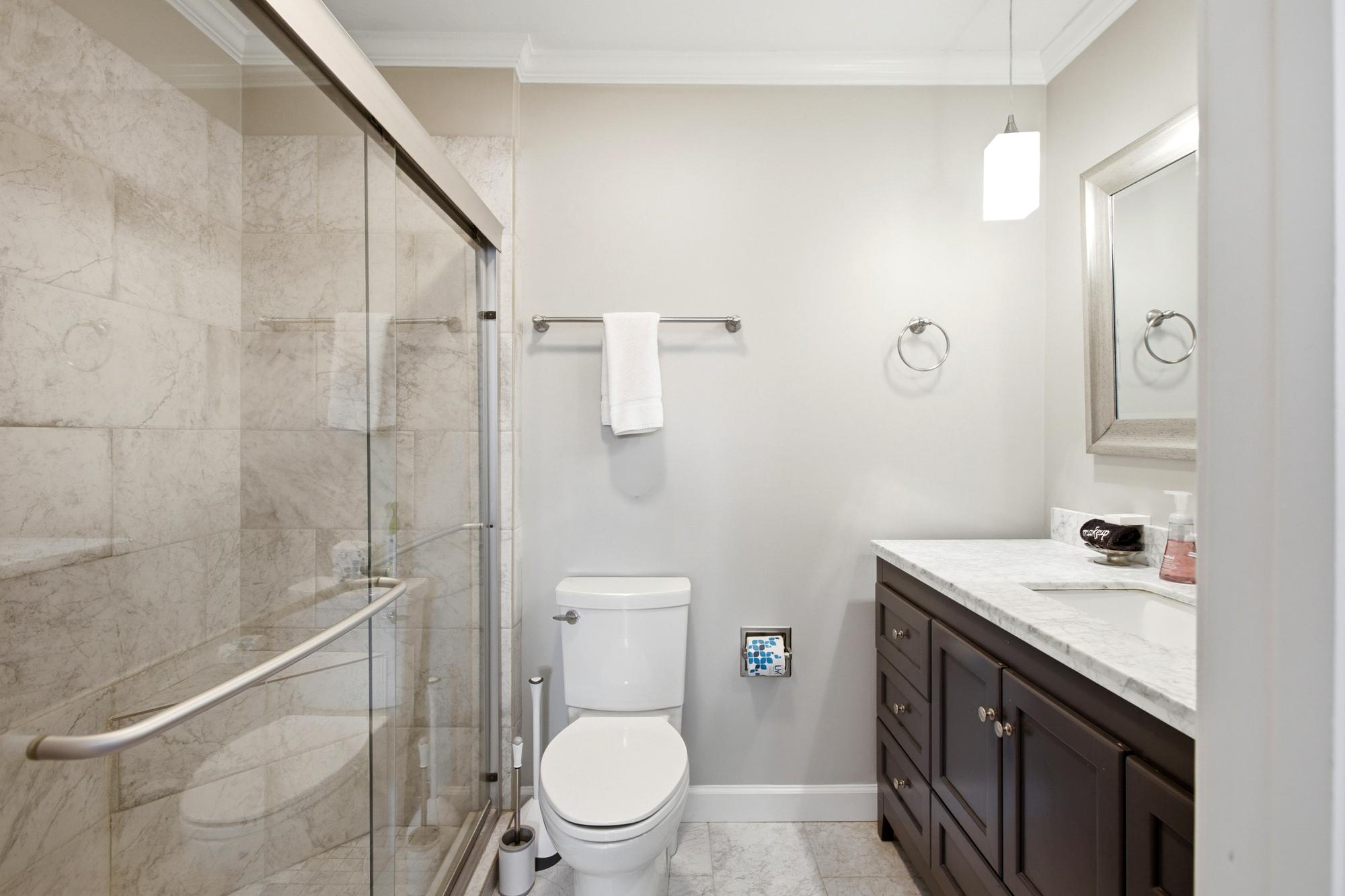 Tiled walls and a spacious glass shower create a spa-like atmosphere in this bright bathroom. Dark wood cabinetry provides a rich contrast to the clean vanities and glowing pendant light.