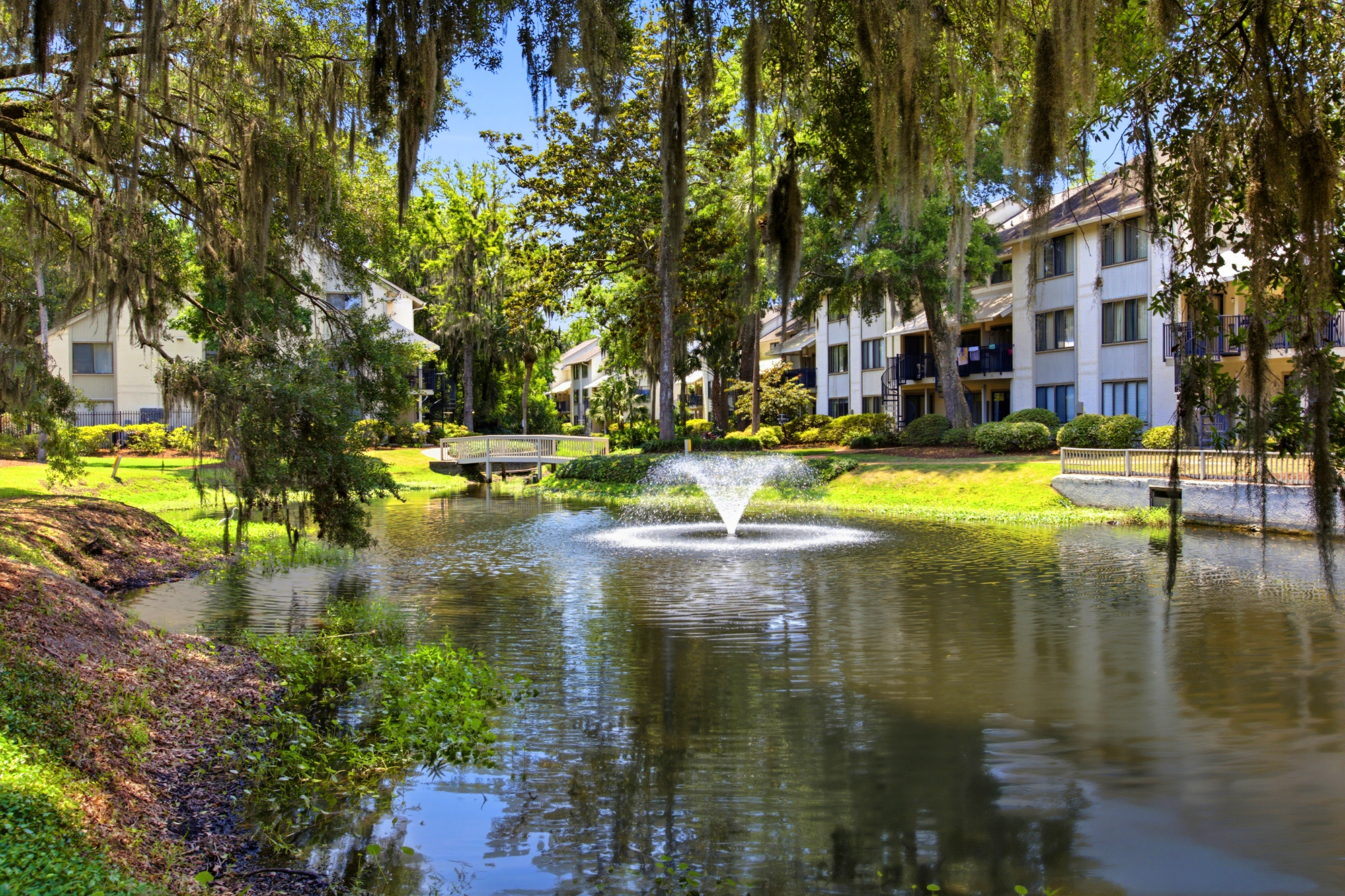 Spanish moss drapes gracefully over a shimmering pond, where a central fountain creates a soothing soundtrack for your stay. Bright afternoon glare illuminates the lush banks and white footbridge, framing a peaceful scene inside Springwood Villas.