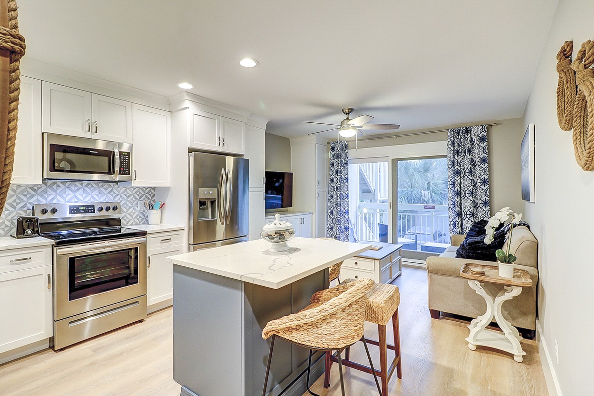 Polished marble counters and honey-toned floors anchor this airy kitchen, where nautical rope accents and patterned linens create a coastal charm. Natural light pours through sliding doors, making every morning meal feel like a seaside escape.