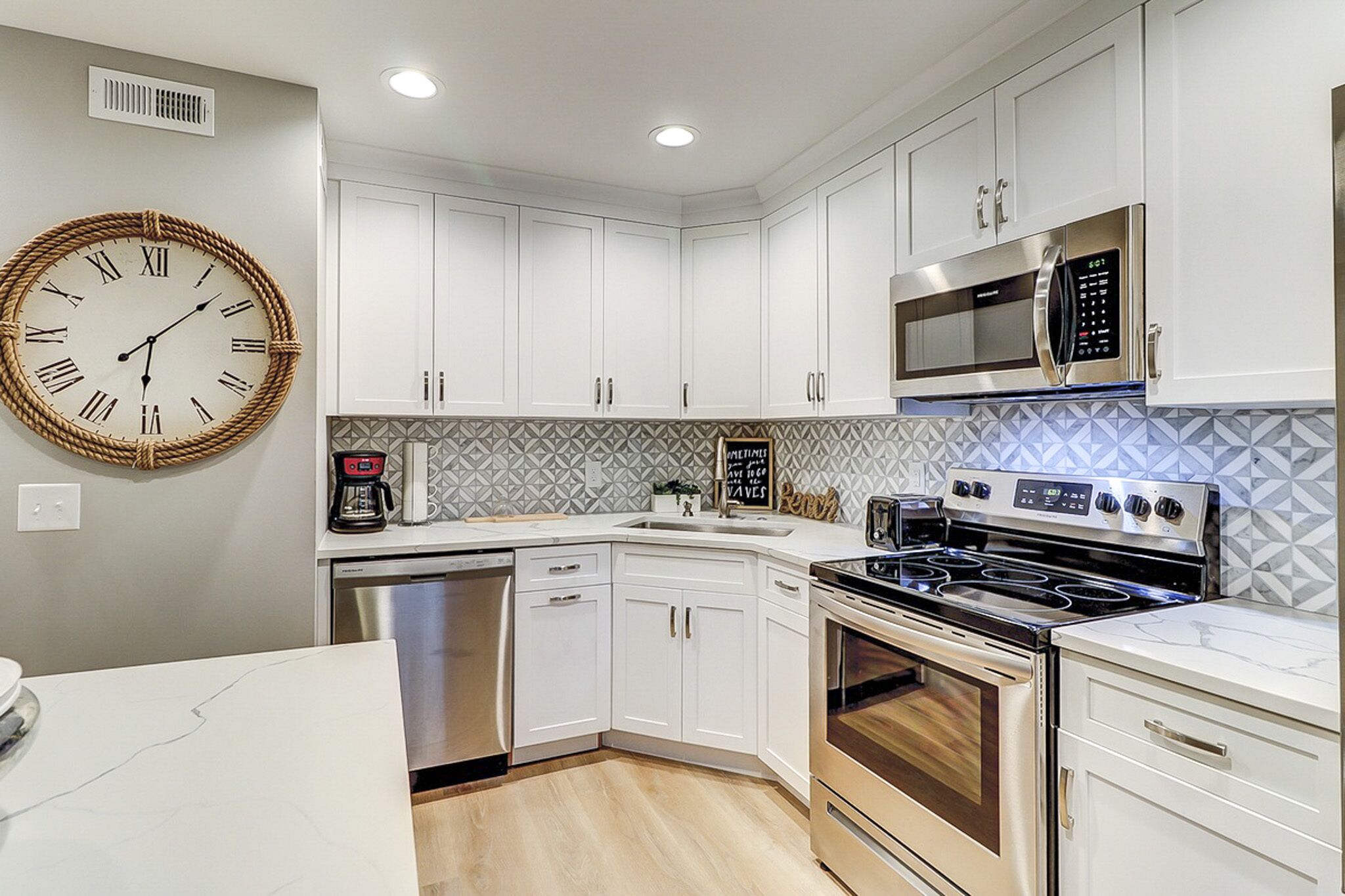 Alabaster cabinetry and a geometric backsplash create a bright, contemporary culinary space. Stainless steel appliances gleam against marble counters, while the oversized rope clock adds a charming nautical touch to this airy kitchen.