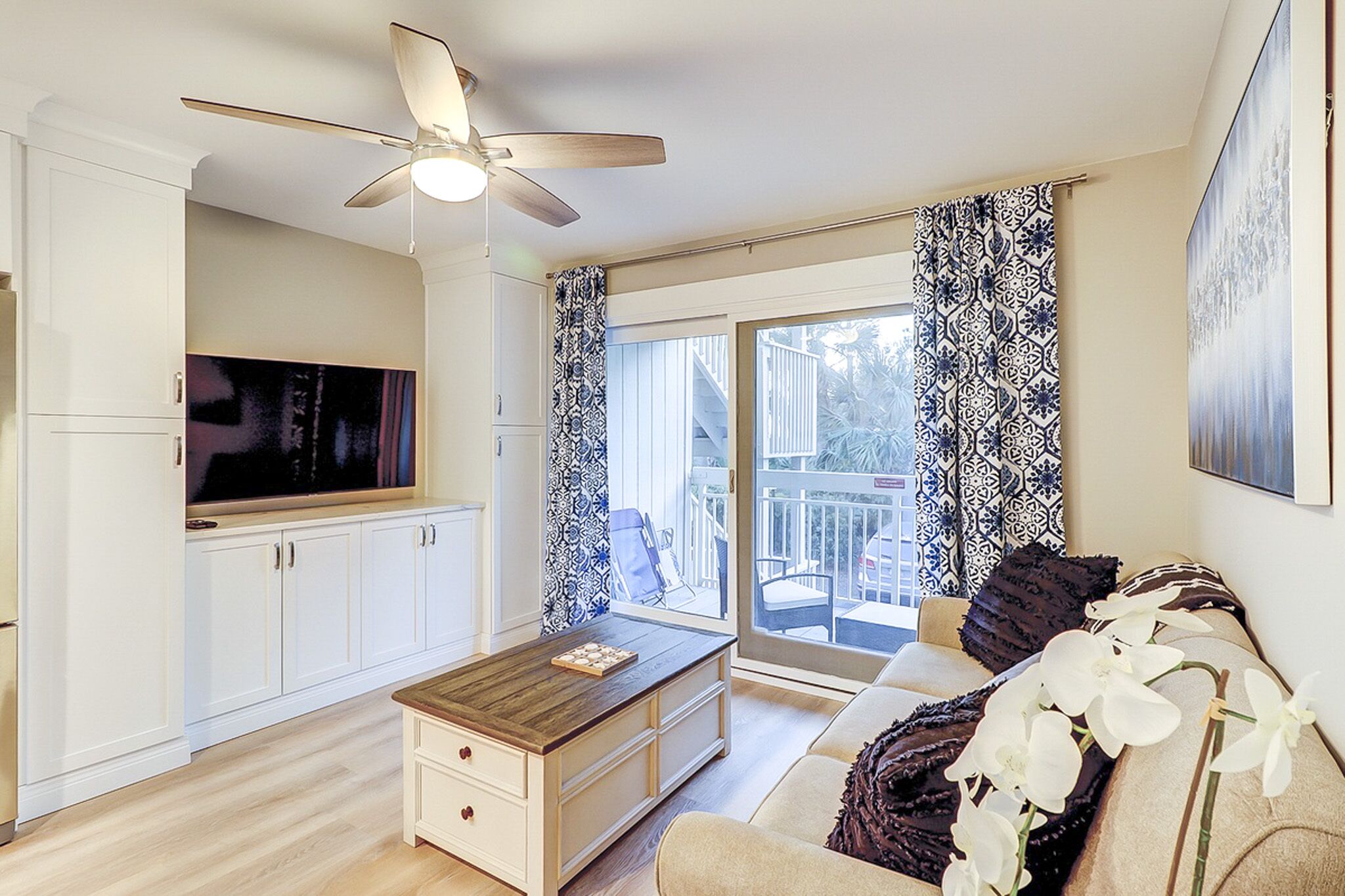 Blue patterned curtains frame glass doors that lead to a private balcony, filling this airy living area with soft daybreak. A warm wood coffee table and creamy built-ins create a calm, organized space for quiet mornings or movie nights.