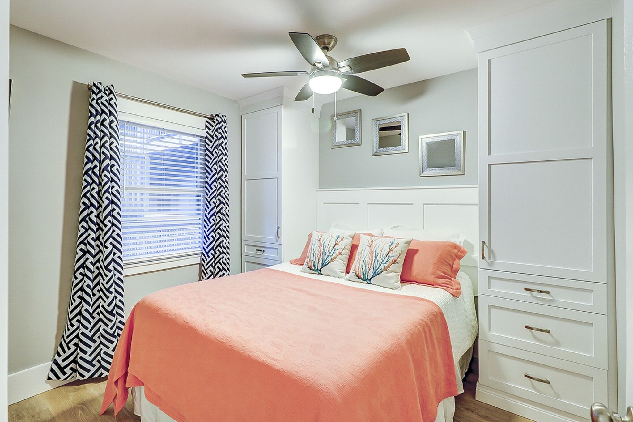 Coral linens and sea-inspired pillows bring a splash of warmth to this airy bedroom. Built-in white cabinetry provides ample storage, while soft natural light filters through patterned curtains for a peaceful morning glow.