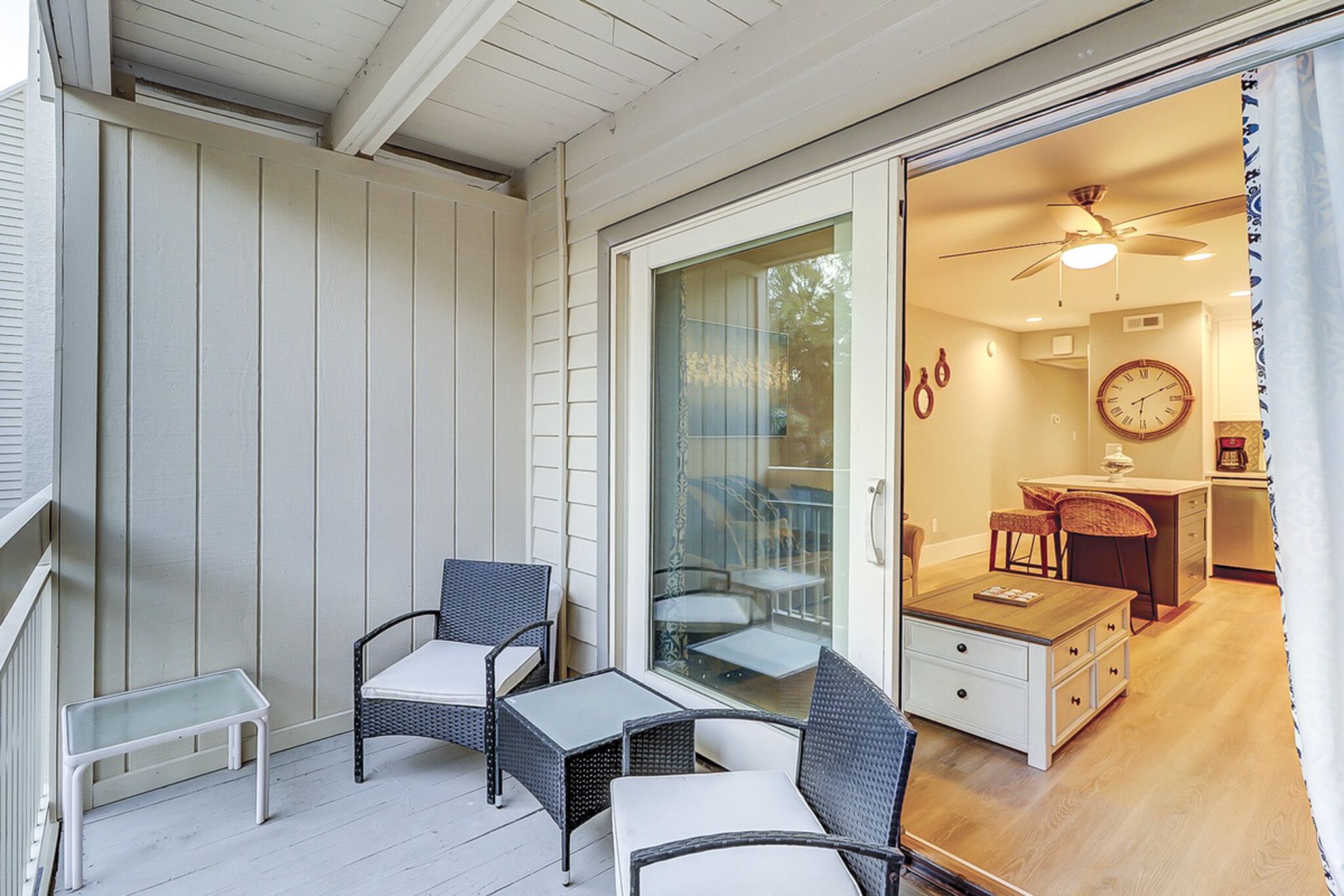 Patio chairs with plush cushions offer a private nook for coffee, separated from the glowing indoor living area by wide glass doors. Warm golden light spills across the wood floors, creating a seamless flow between the fresh air and cozy interior.