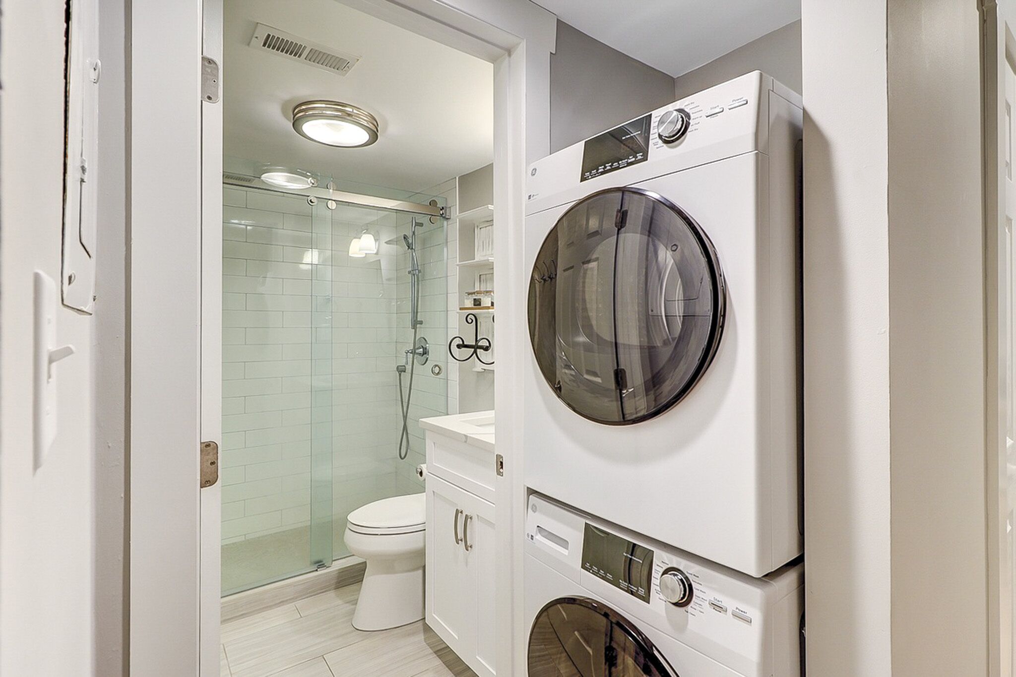 Modern convenience meets sleek design in this efficient utility space, featuring a stacked washer and dryer next to a bright bathroom. Glass shower doors and subway tiles reflect the soft glow for a clean, refreshing atmosphere throughout.