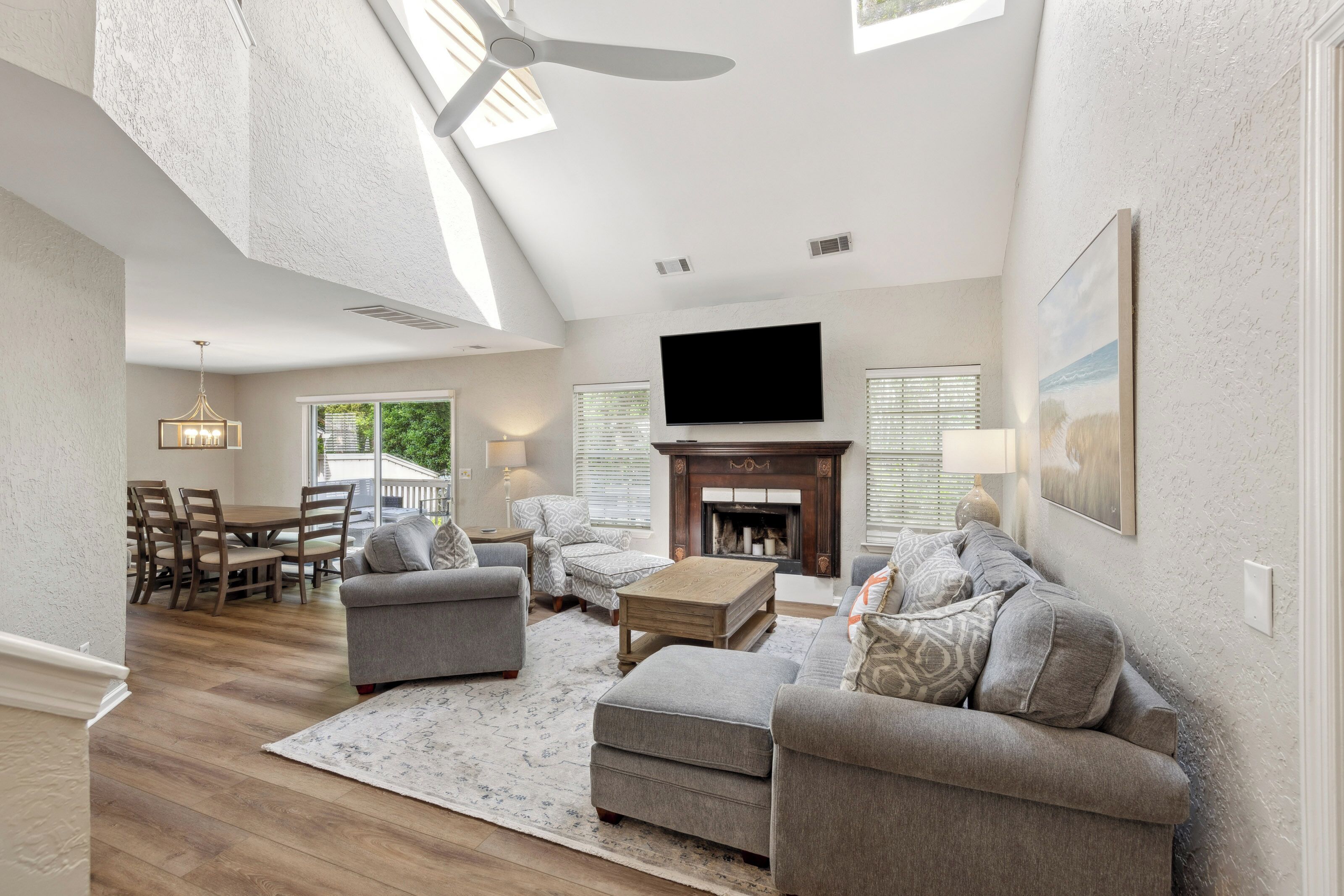 Plush grey seating and a warm wood fireplace anchor this airy living room, where vaulted ceilings and skylights invite a soft afternoon glow. Polished floors and coastal art create a serene, open space perfect for quiet mornings or movie nights.