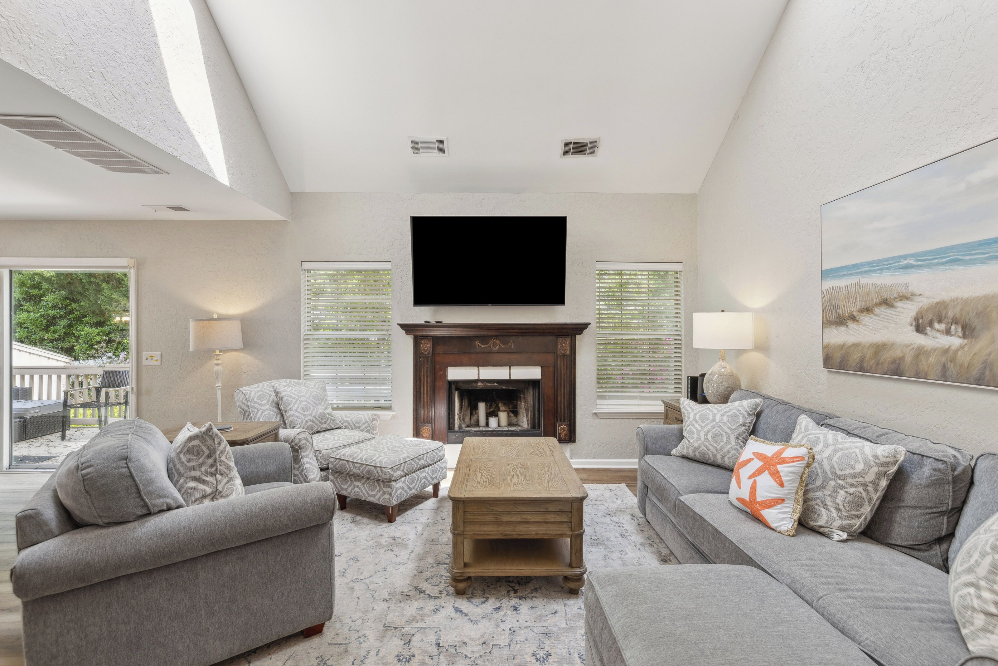 Vaulted ceilings and a deep wood fireplace anchor this airy living room, where soft grey linens meet coastal accents. An afternoon glow pours through large windows, creating a serene spot for quiet mornings or movie nights by the hearth.