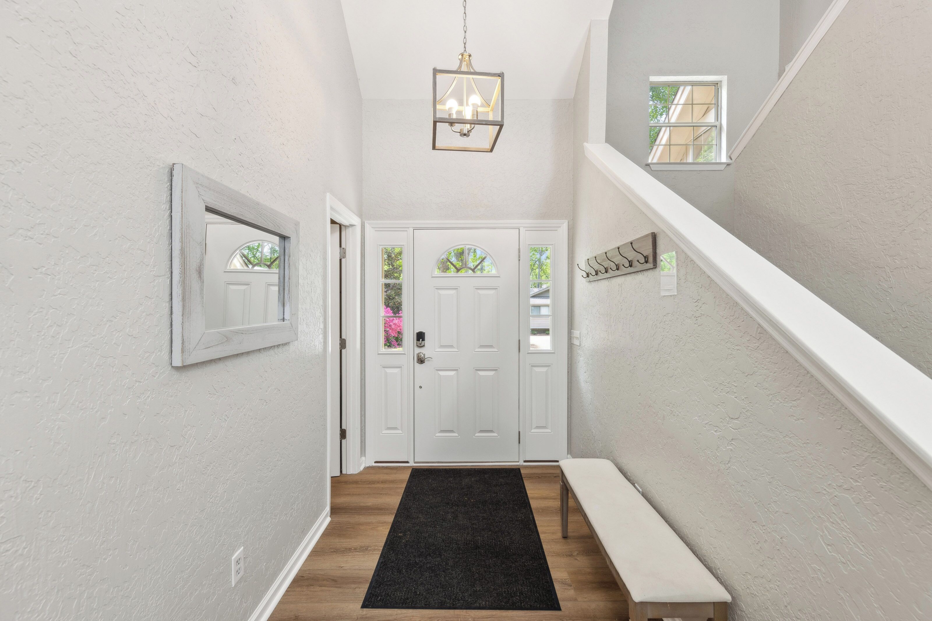 Bright white walls and warm wood floors create a crisp, airy entrance. A modern lantern glows above, casting soft light over the clean lines and welcoming bench of this sun-drenched foyer.