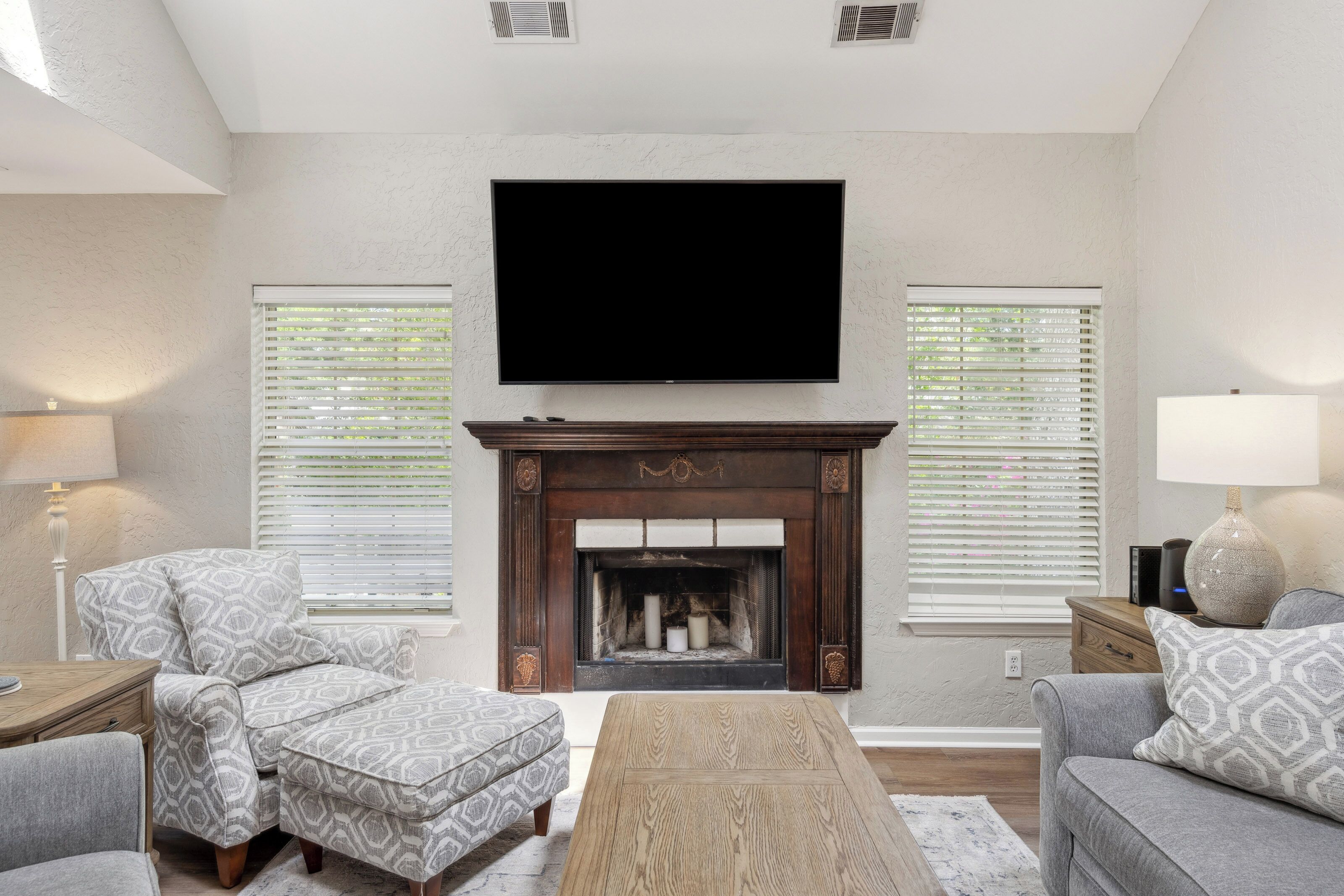 Ornate wood mantelpiece accents anchor this cozy living room, where soft grey patterns and a sun-drenched atmosphere invite you to linger. Afternoon glow filters through the blinds, highlighting the rich textures and crisp, high ceilings.