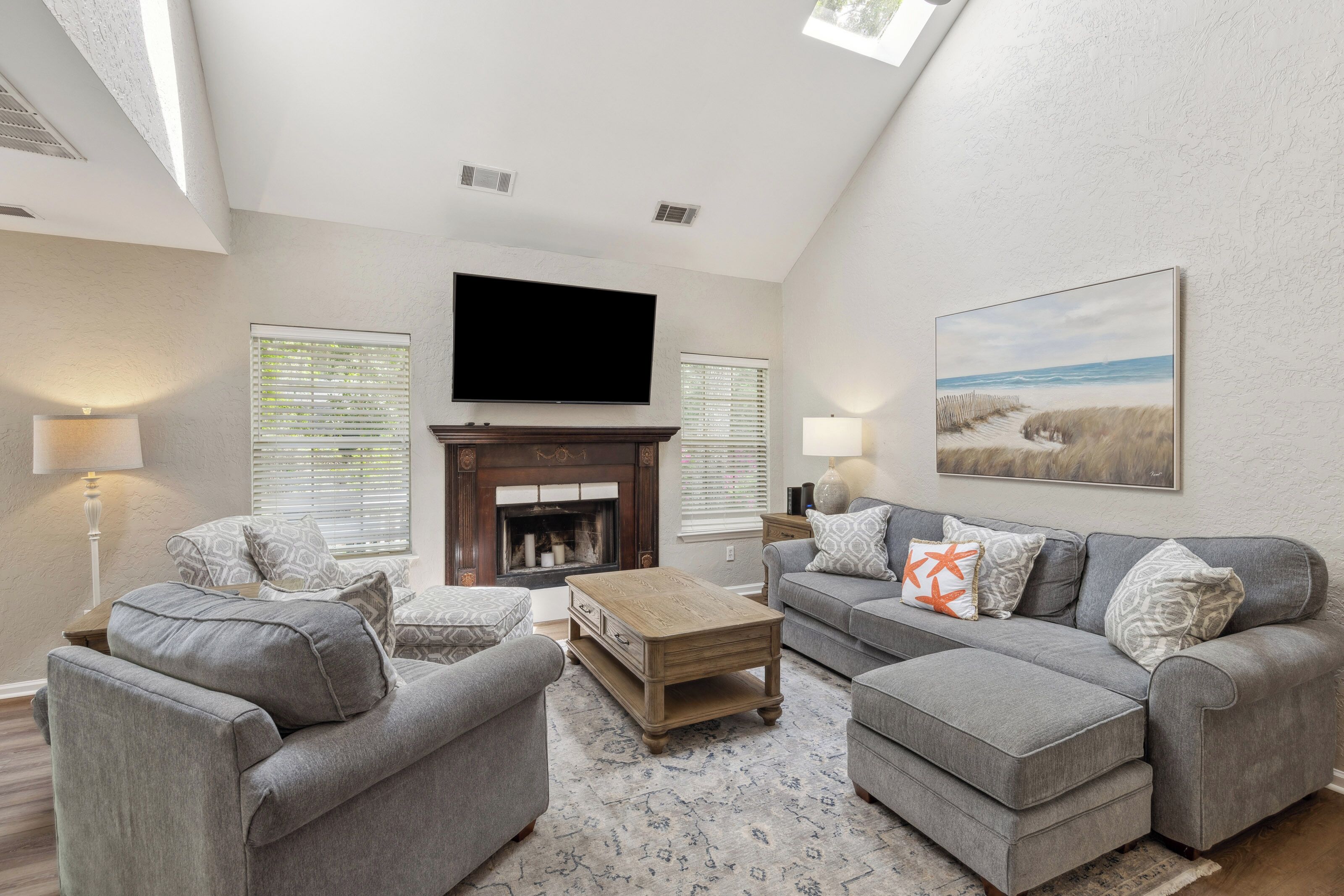 Warm rays pour through the skylight, illuminating a coastal-inspired lounge where soft textiles and a rich wood hearth create an inviting atmosphere. Sink into the generous seating for a movie night under the airy, high ceilings.