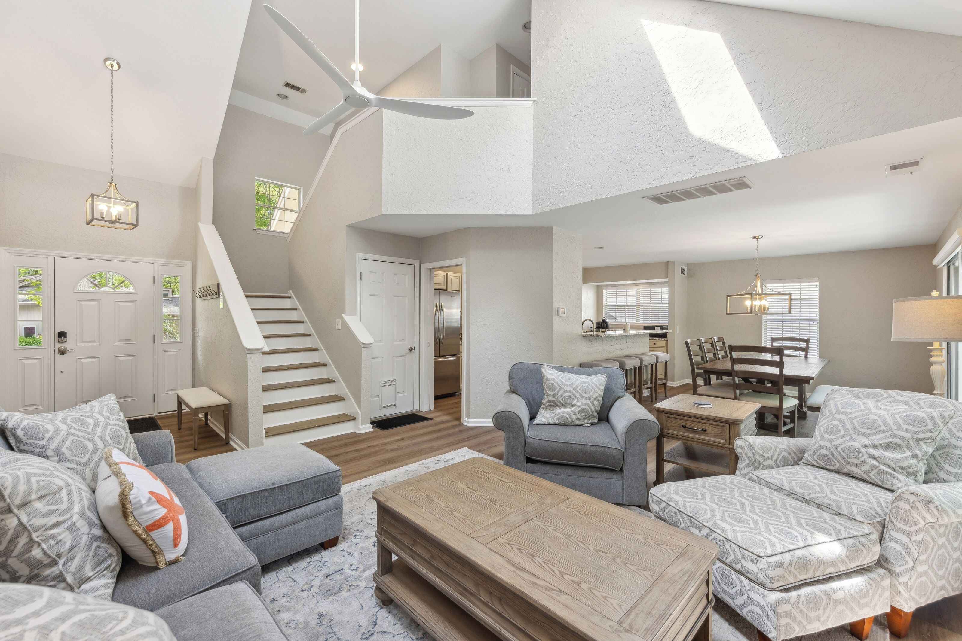 Airy double-height ceilings invite a soft afternoon glow into this open living space, where textured wood and cool grey upholstery offer a calm spot to gather.