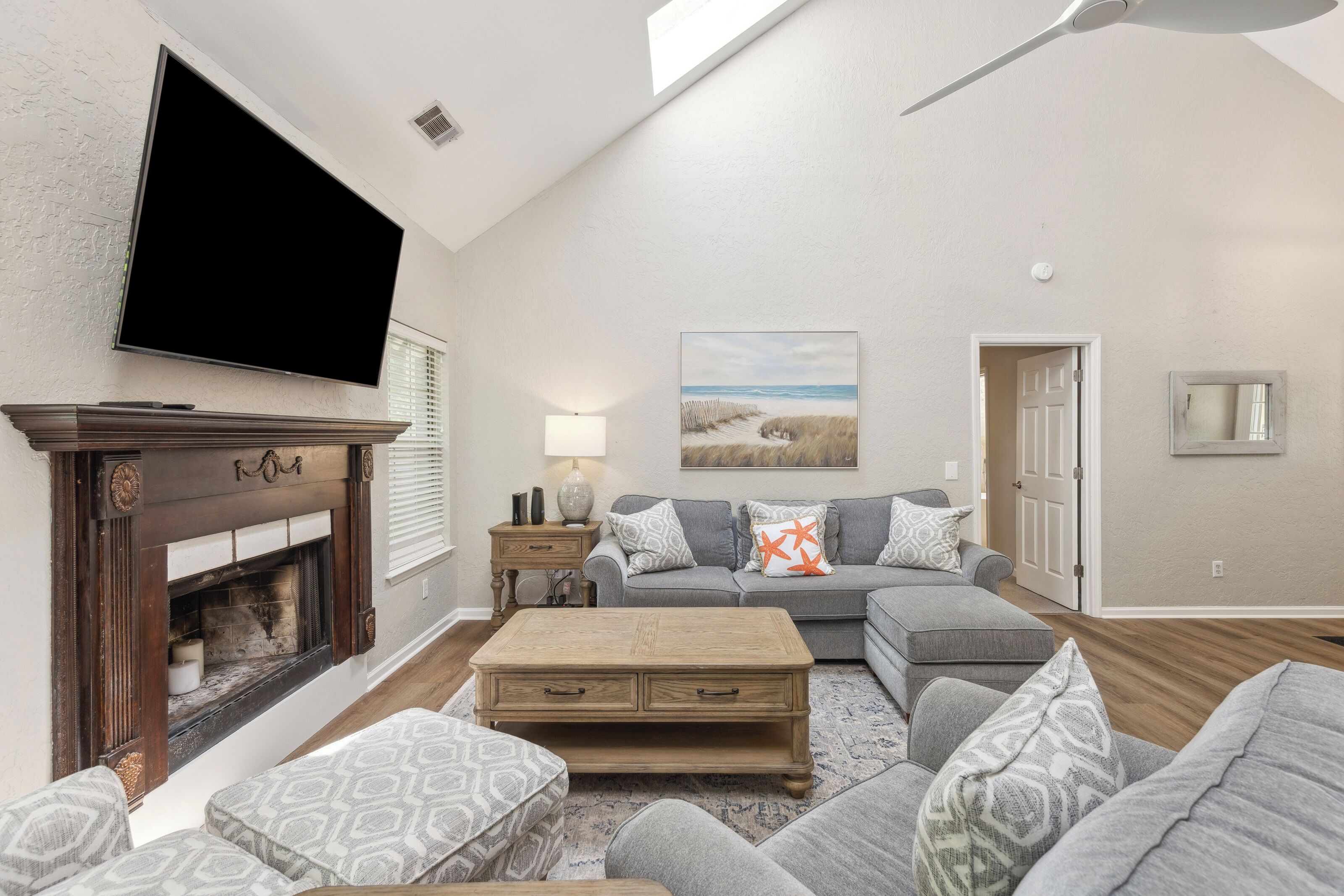 Polished wood and soft grey fabrics anchor this airy living room, where a skylight pours an afternoon glow over the seating area. The ornate fireplace adds a touch of classic charm to a space designed for quiet evenings and coastal comfort.