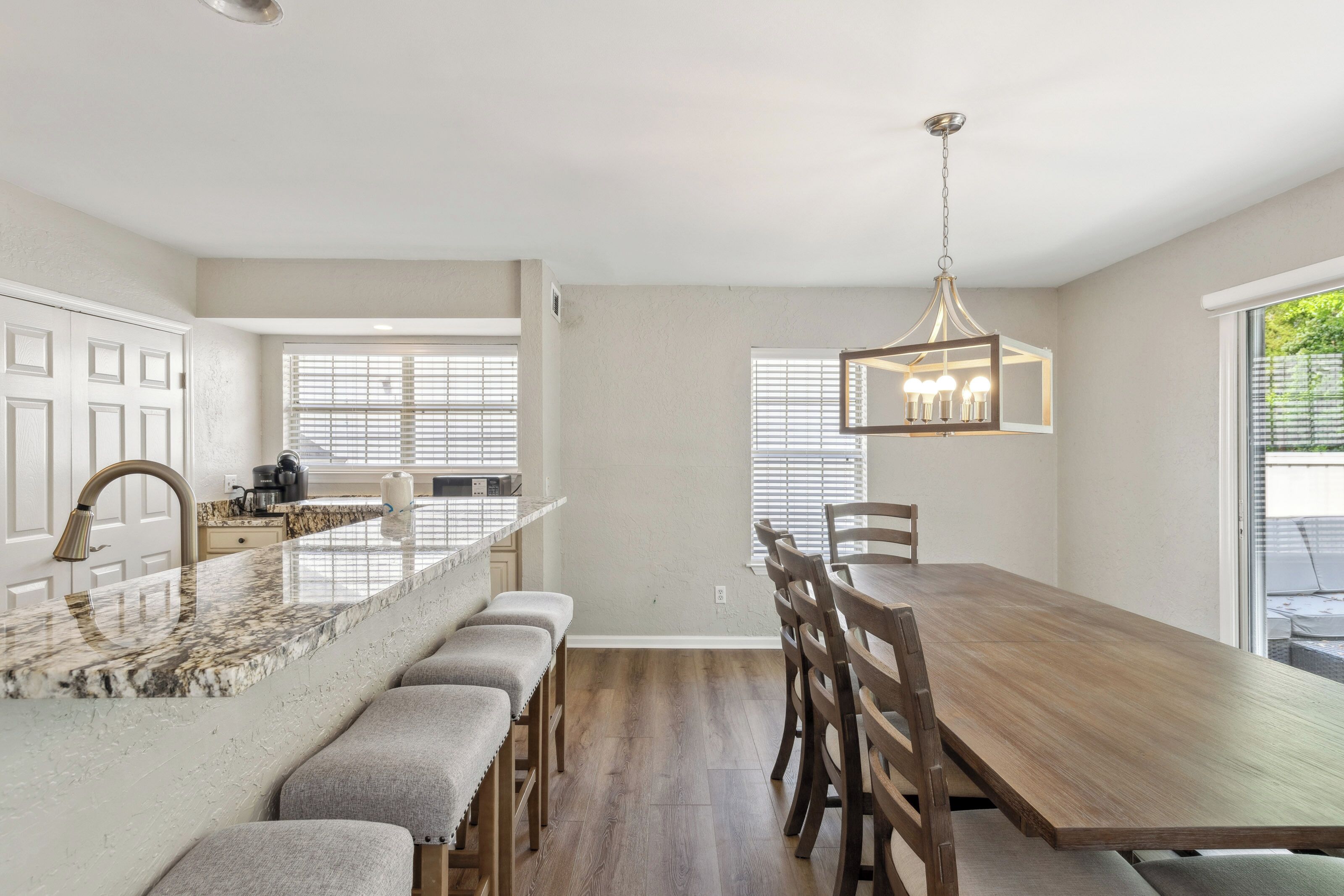 Speckled stone counters and soft grey seating create a sophisticated spot for morning coffee. This open dining area glows with afternoon rays, offering a sturdy wooden table and a modern chandelier for memorable shared meals.