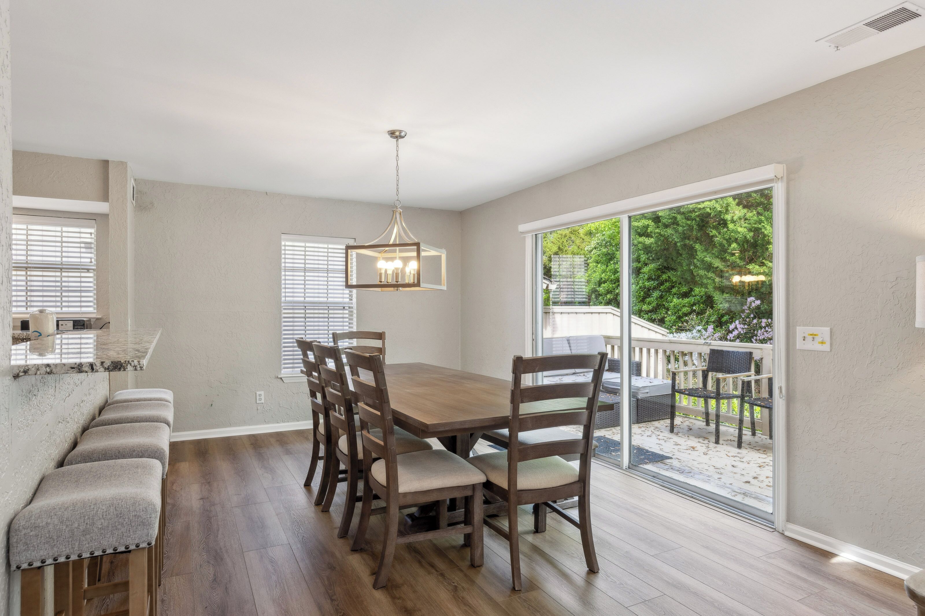 Timber textures and soft upholstery create a grounded, cozy dining atmosphere. Large glass doors bridge the gap between the indoor table and the leafy deck, inviting a gentle afternoon glow to fill the open room.