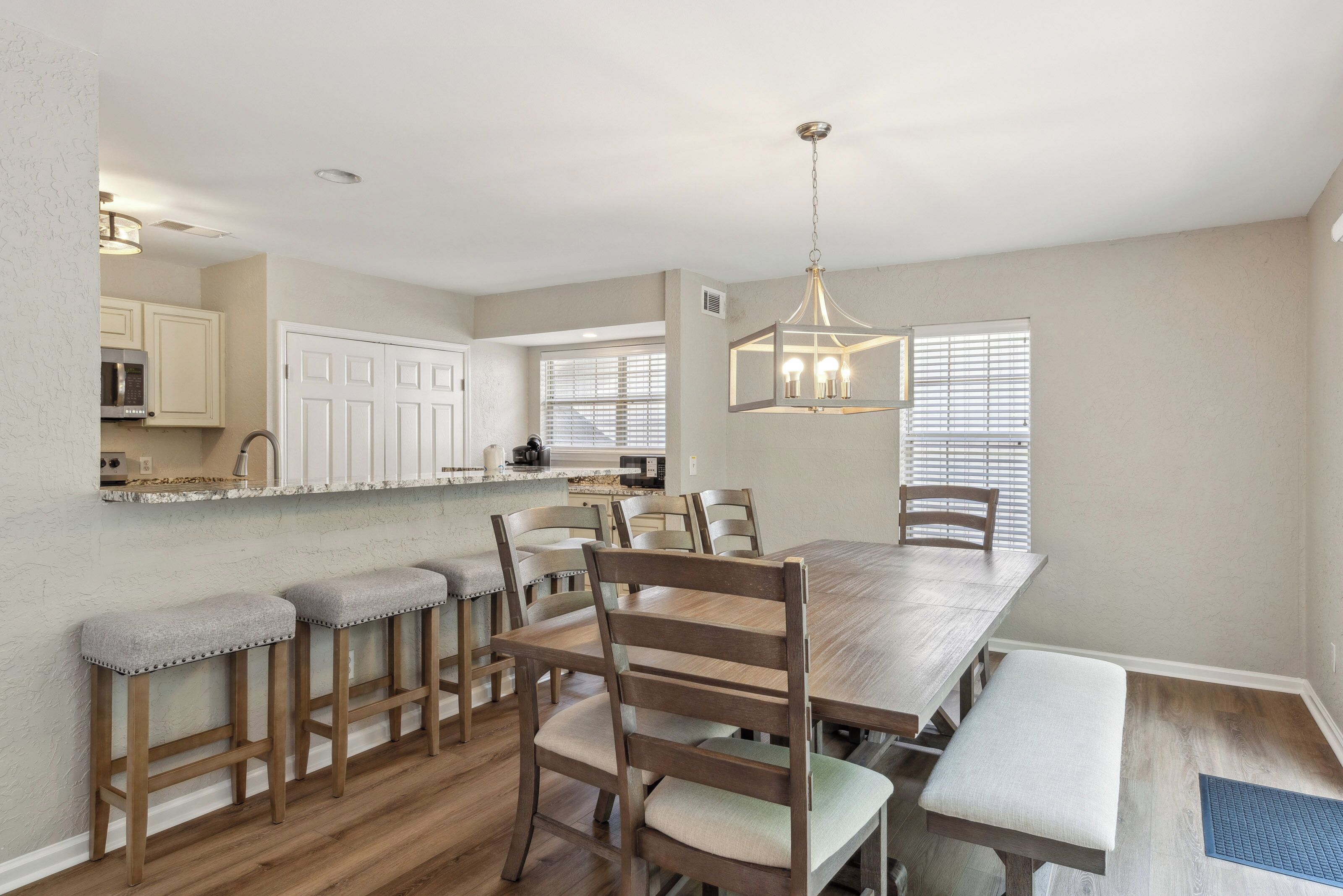 Granite counters and a sturdy wooden dining table anchor this open heart of the home, where soft afternoon glow meets crisp, neutral tones. Padded seating and a modern lantern fixture invite slow mornings and shared meals in a clean, calm space.
