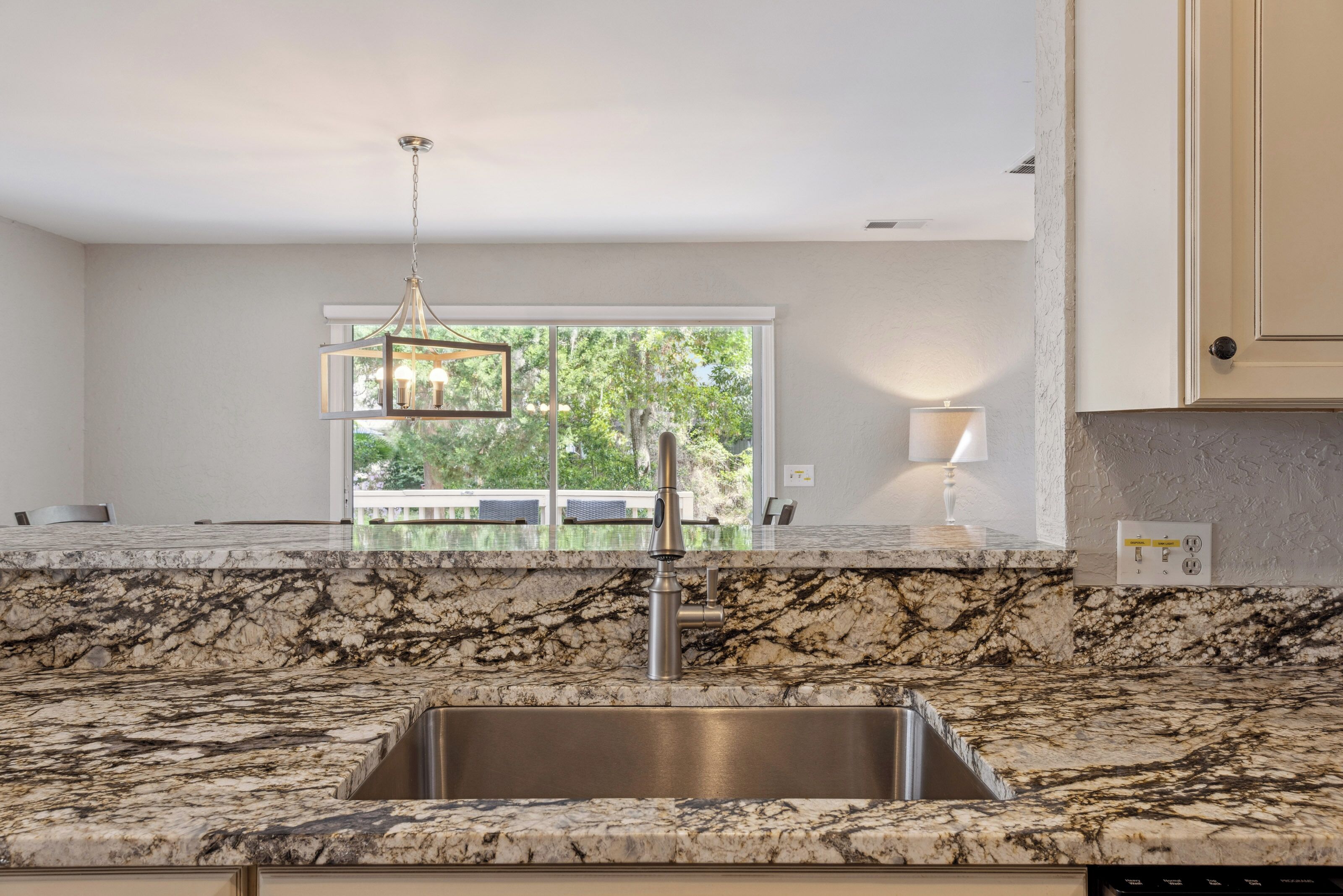 Earthy veined countertops and a deep stainless sink create a sophisticated space for meal prep. Beyond the breakfast bar, soft lamp glows and leafy views through the sliding glass door offer a peaceful backdrop for your morning coffee.